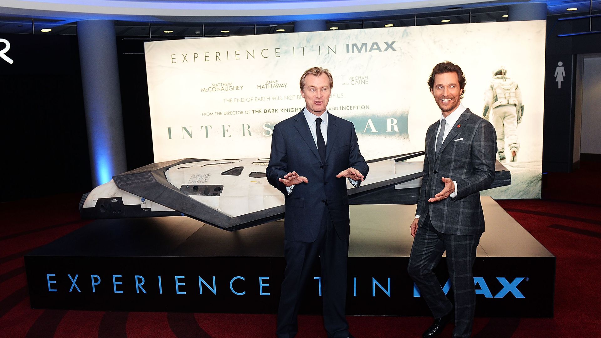 Two men in suits pose in front of a large Interstellar IMAX banner, with a life-size spaceship model and an astronaut image; blue ceiling lights above.