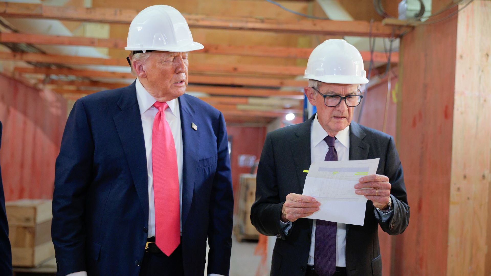 Two men in suits and white hard hats in a wooden construction area, one with a red tie looking at the other who is reading a document with highlighted text.