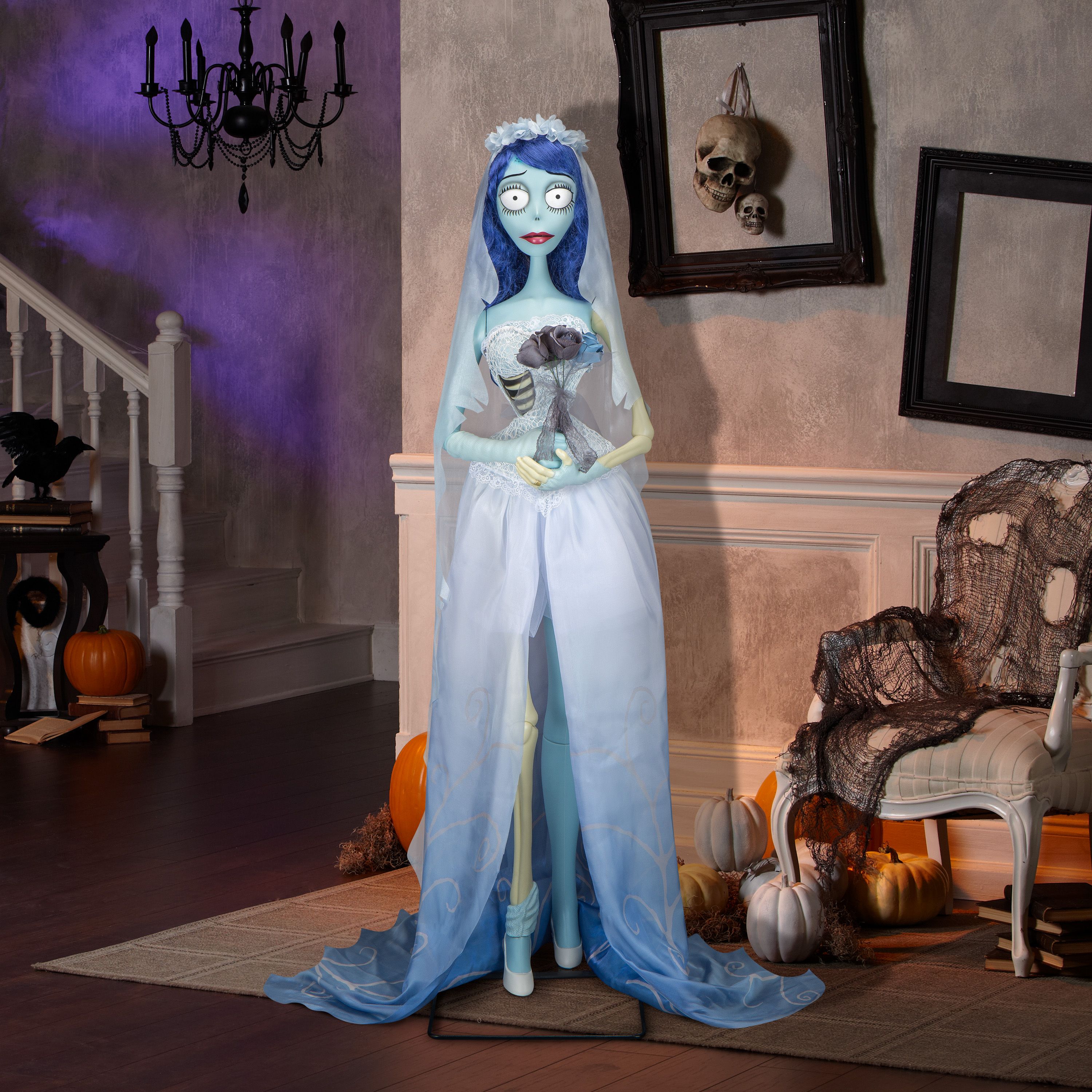 A bride figurine with blue hair and white veil in wedding dress holding gray flowers, standing in Halloween-themed room with pumpkins, skulls, and black chandelier.