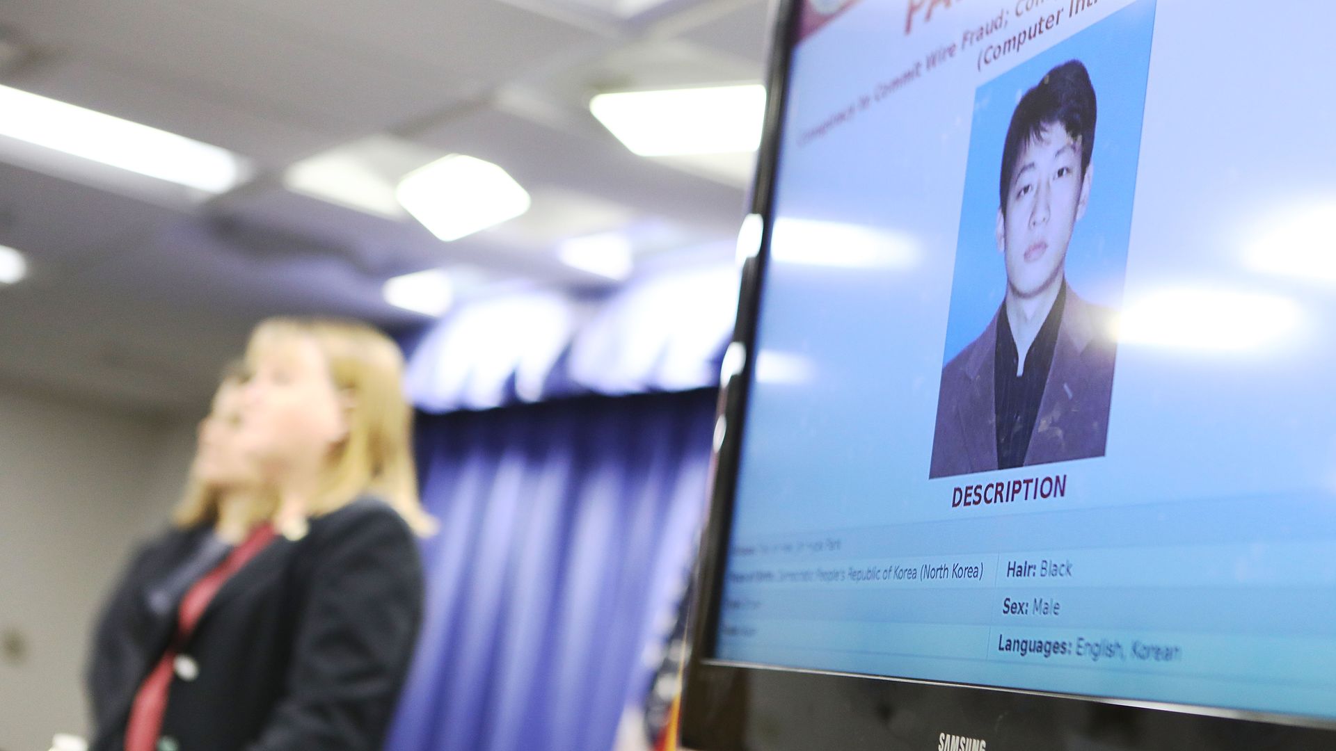 Justice Department officials in the background of a monitor displaying a North Korean man.