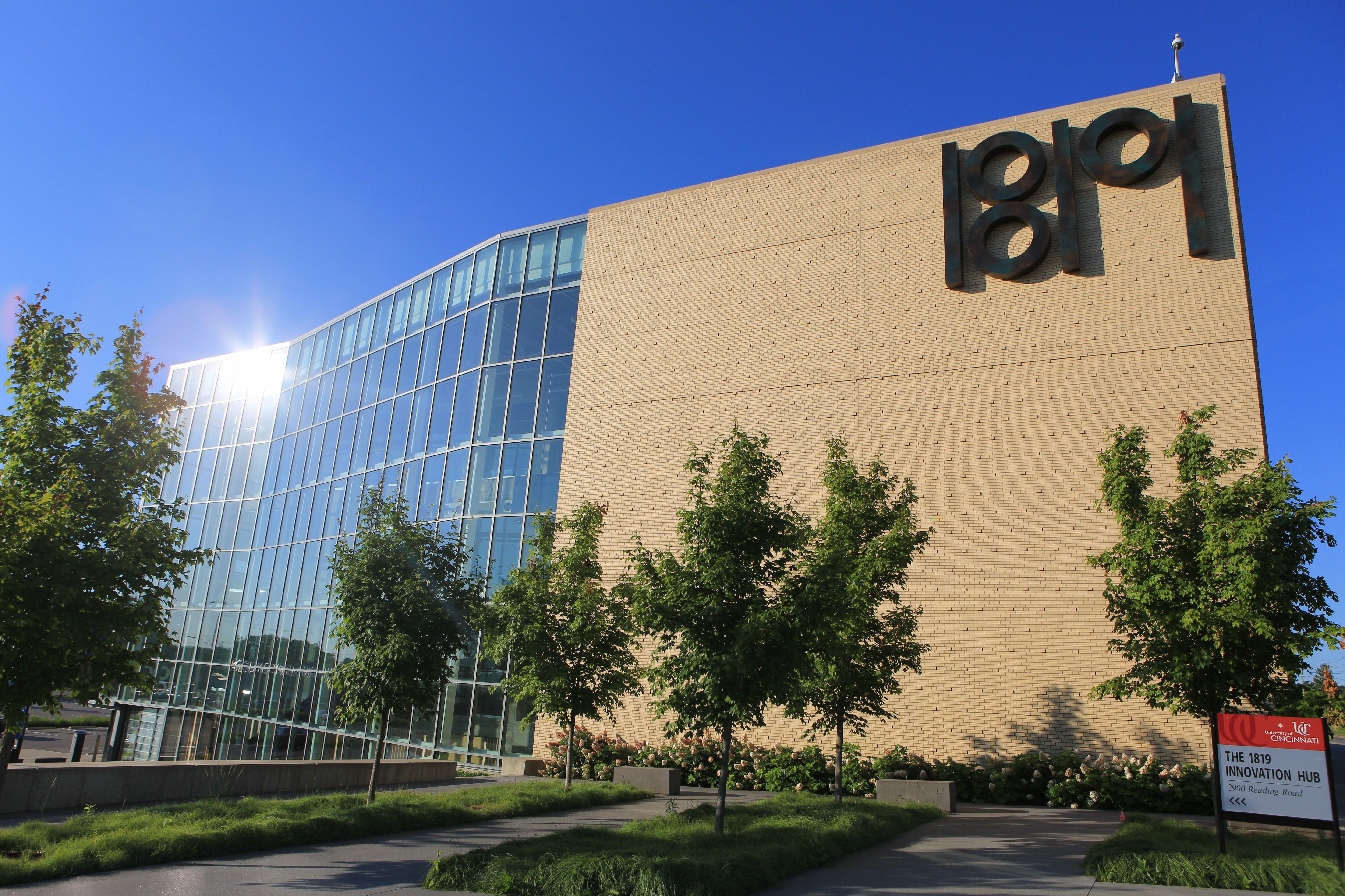 Sunlit modern building with glass on the left and beige brick on the right, large black LIB letters on the wall. Trees in front, blue sky, and a sign for "The 1819 Innovation Hub" at bottom right.