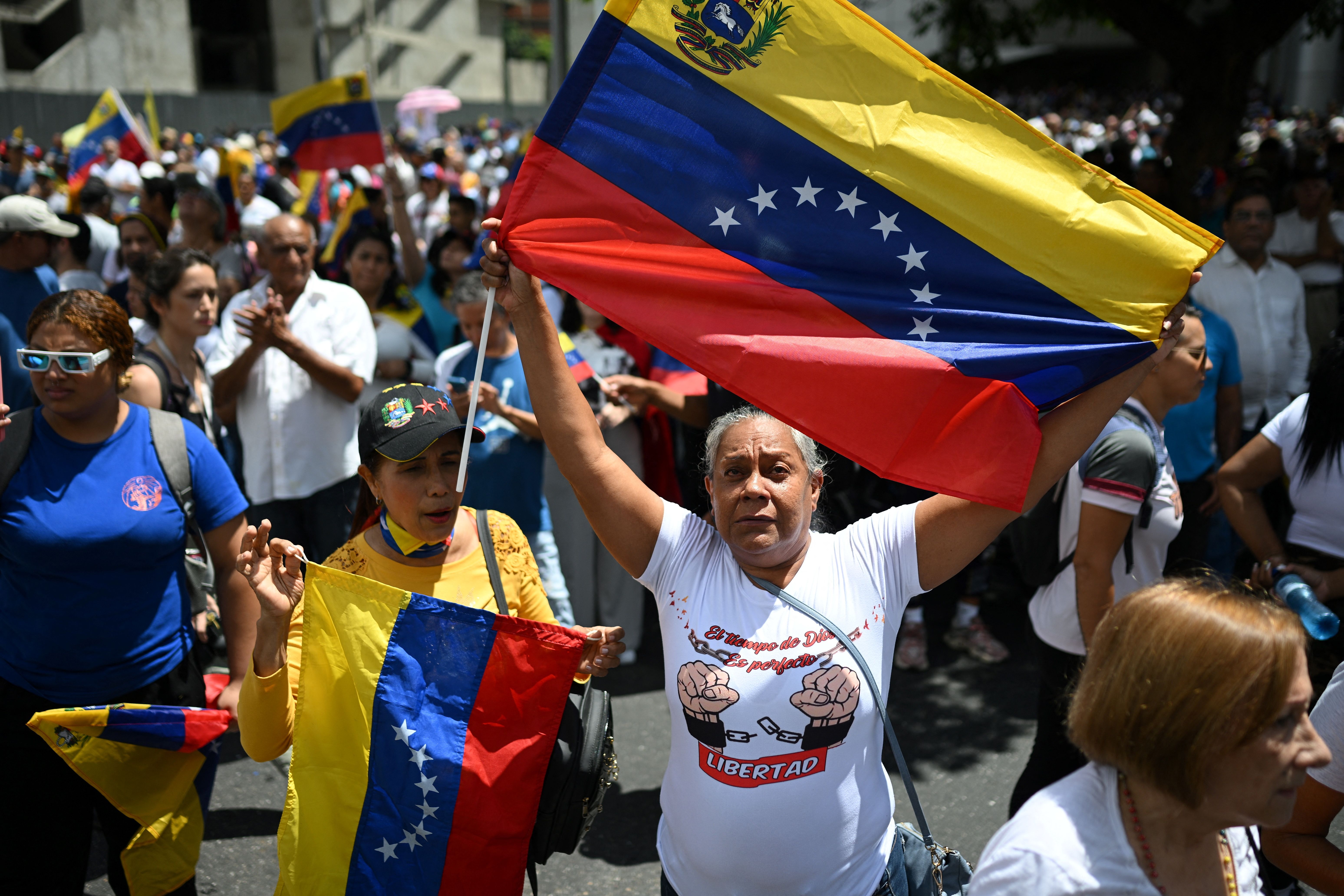A woman protesting in Caracas, Venezuela, holds a large Venezuelan flag up in the air. People are all around her. 