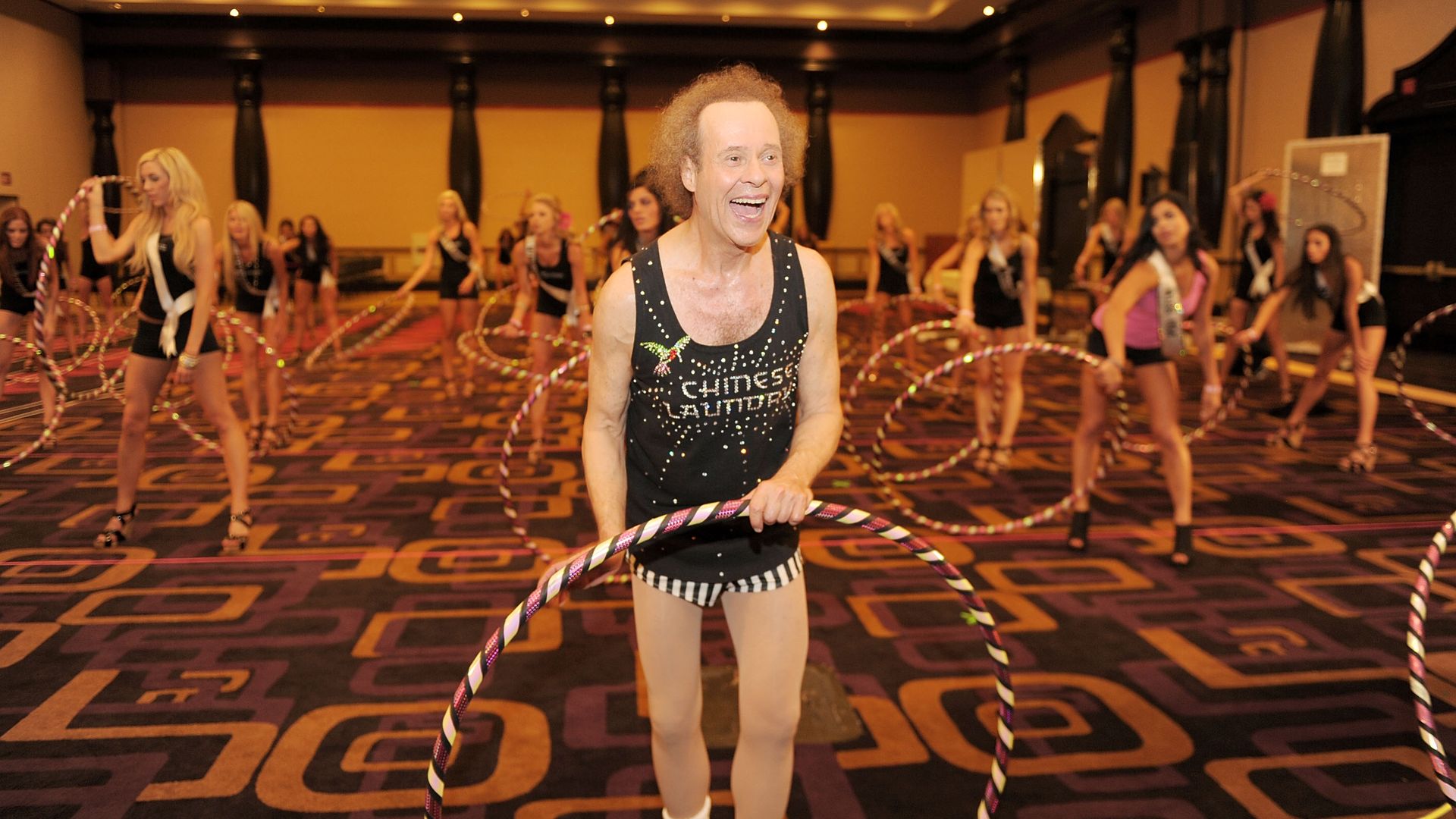 Richard Simmons laughs toward something off camera while holding a hula hoop in a room full of pageant queens hula-hooping.