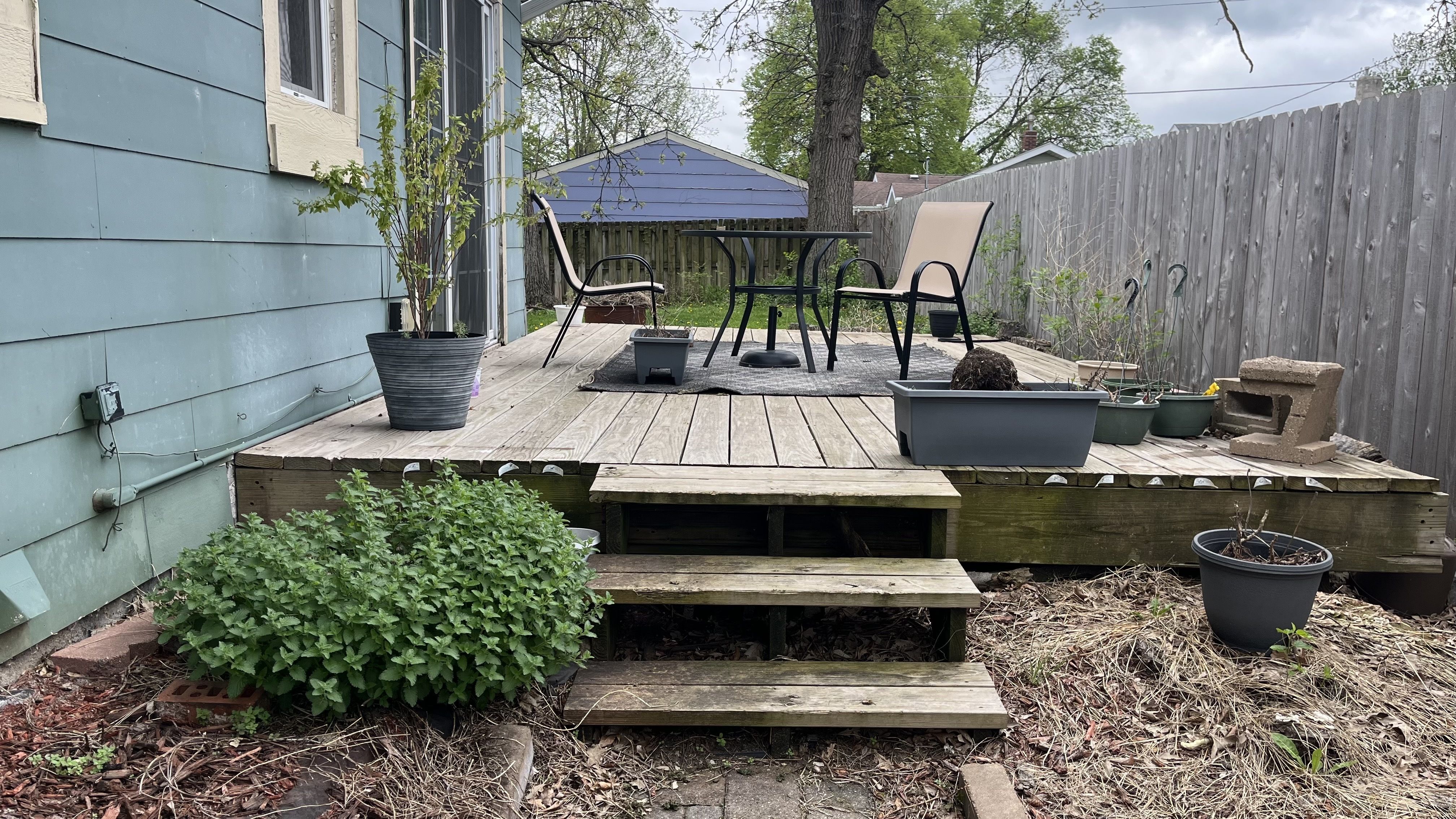 Backyard deck with a black metal table, two beige chairs, and a beige lounge chair. Gray pots with plants, blue house siding on the left, tall wooden fence on the right; overcast sky.