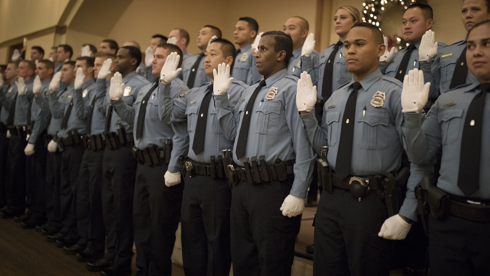 Uniformed police officer trainees stand in formation indoors, raising their right hands in oath. They wear light blue shirts, black ties, white gloves, badges, and duty belts.