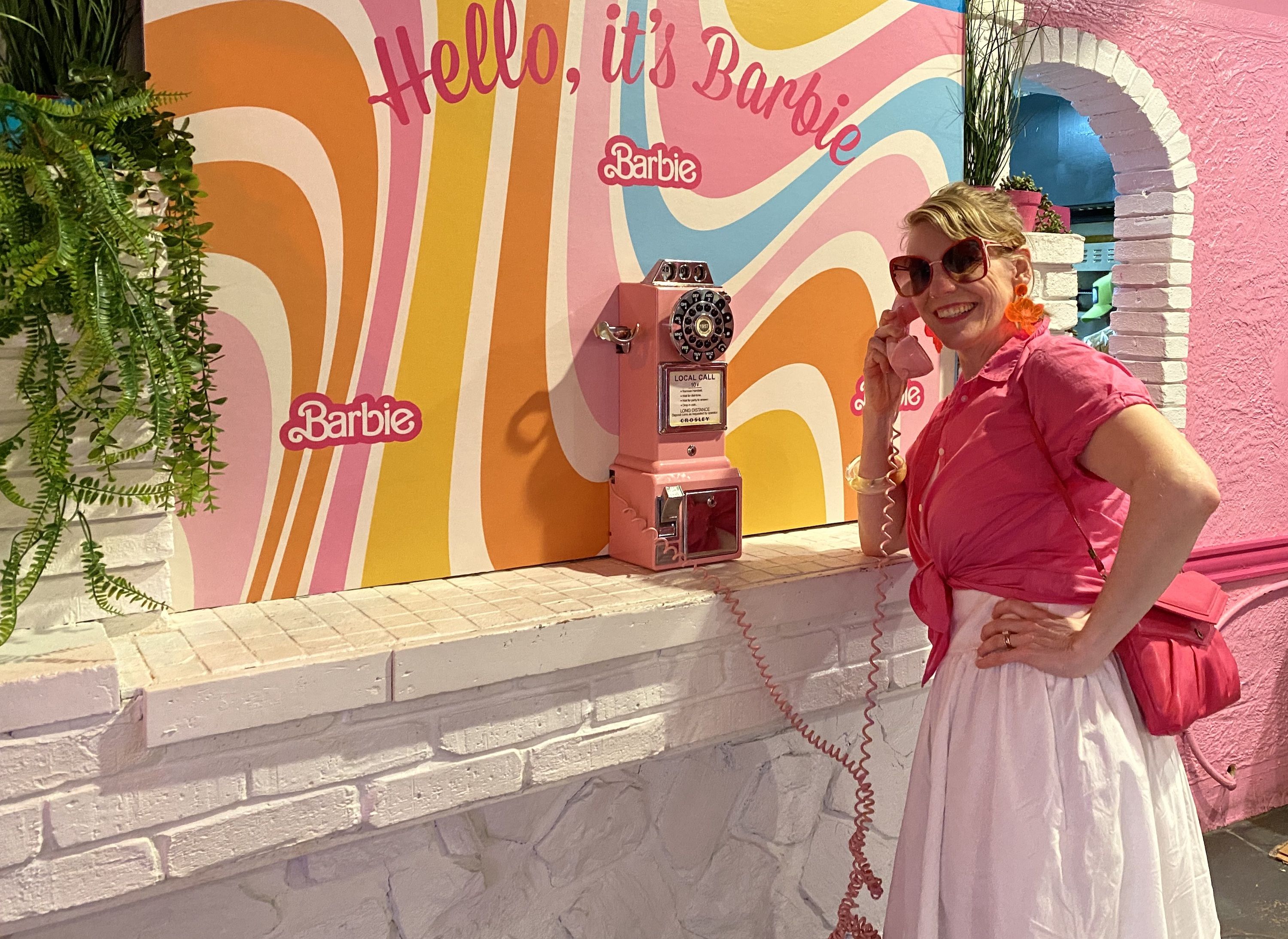 Multi color background that reads "Hello, it's Barbie" with a pink rotary phone which is held by woman in pink shirt and pink sunglasses.