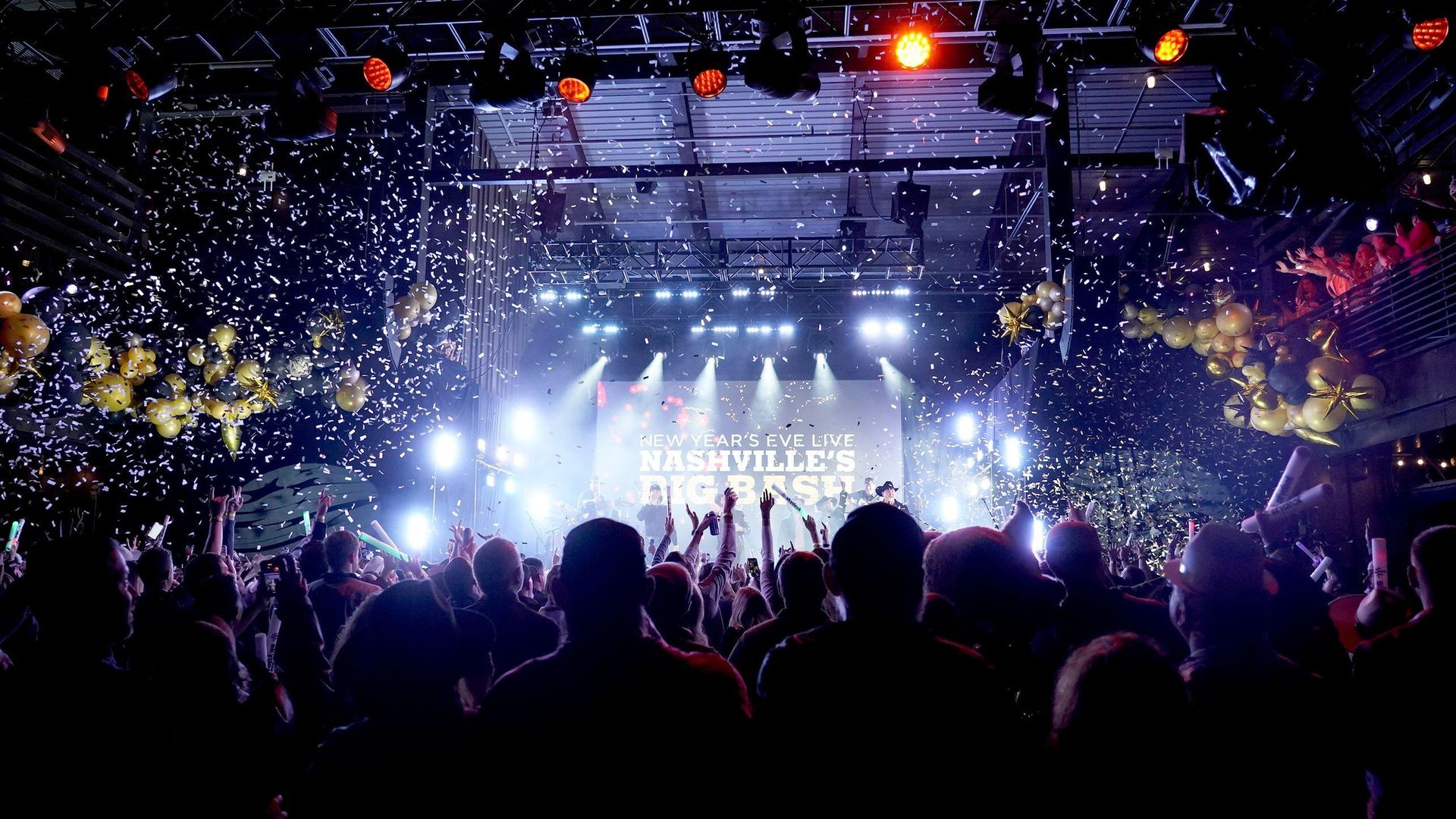 A crowd faces the stage at a music concert as confetti falls.