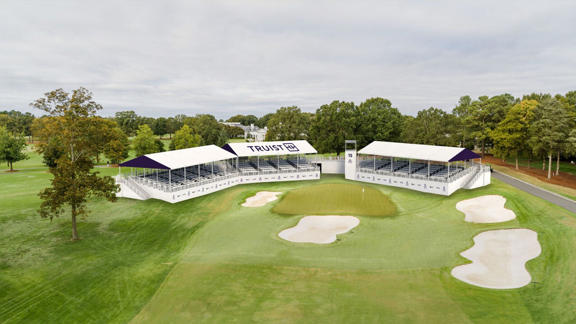 Golf course green with sand bunkers and temporary grandstands labeled "TRUIST" set up for spectators under cloudy sky, surrounded by trees and a distant house.