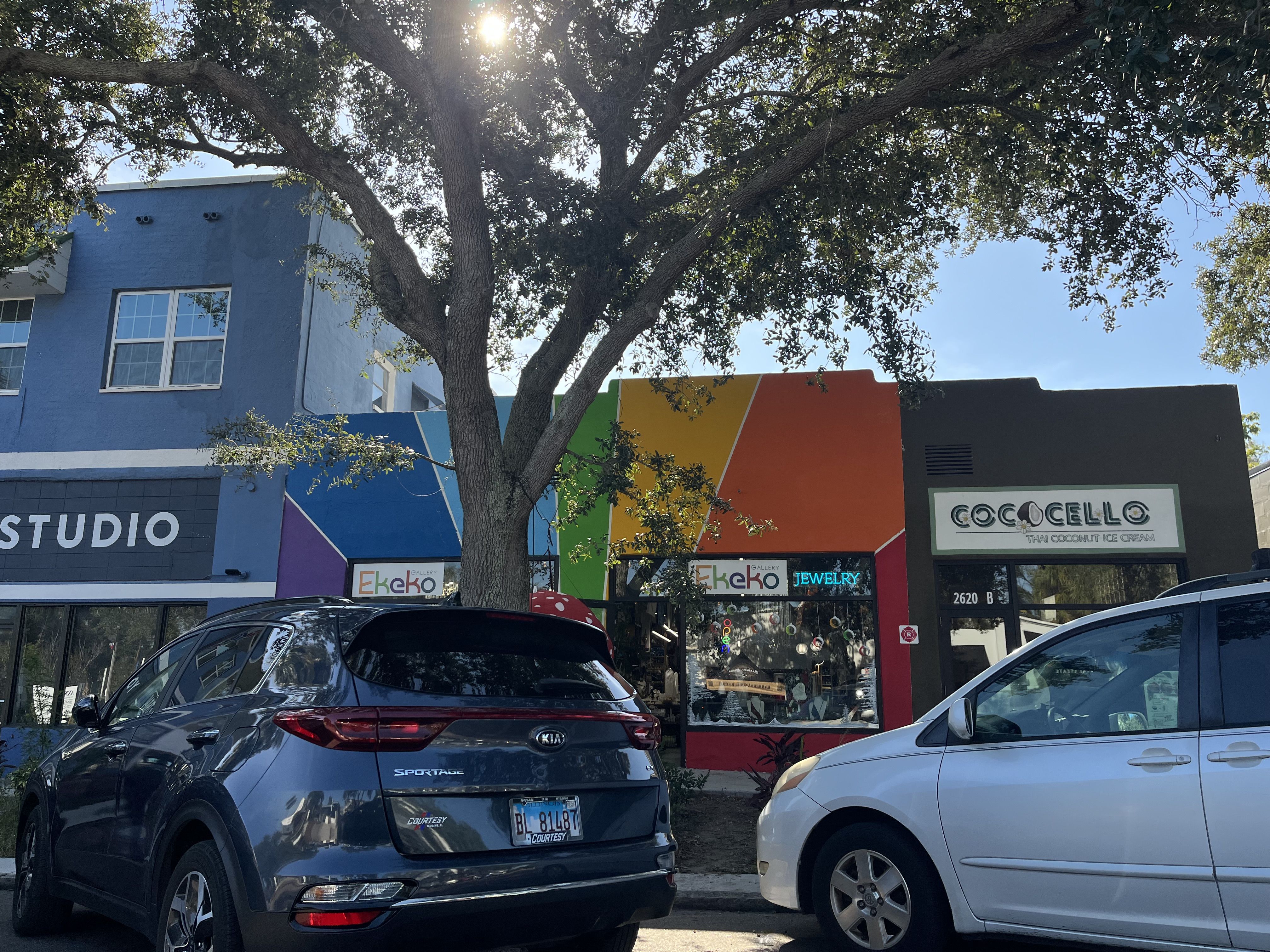 Cars parked in front of colorful storefronts including a blue studio, a rainbow-colored art gallery named Ekeko, and a dark gray Thai coconut ice cream shop named CoccoCello under a tree with sunlight.