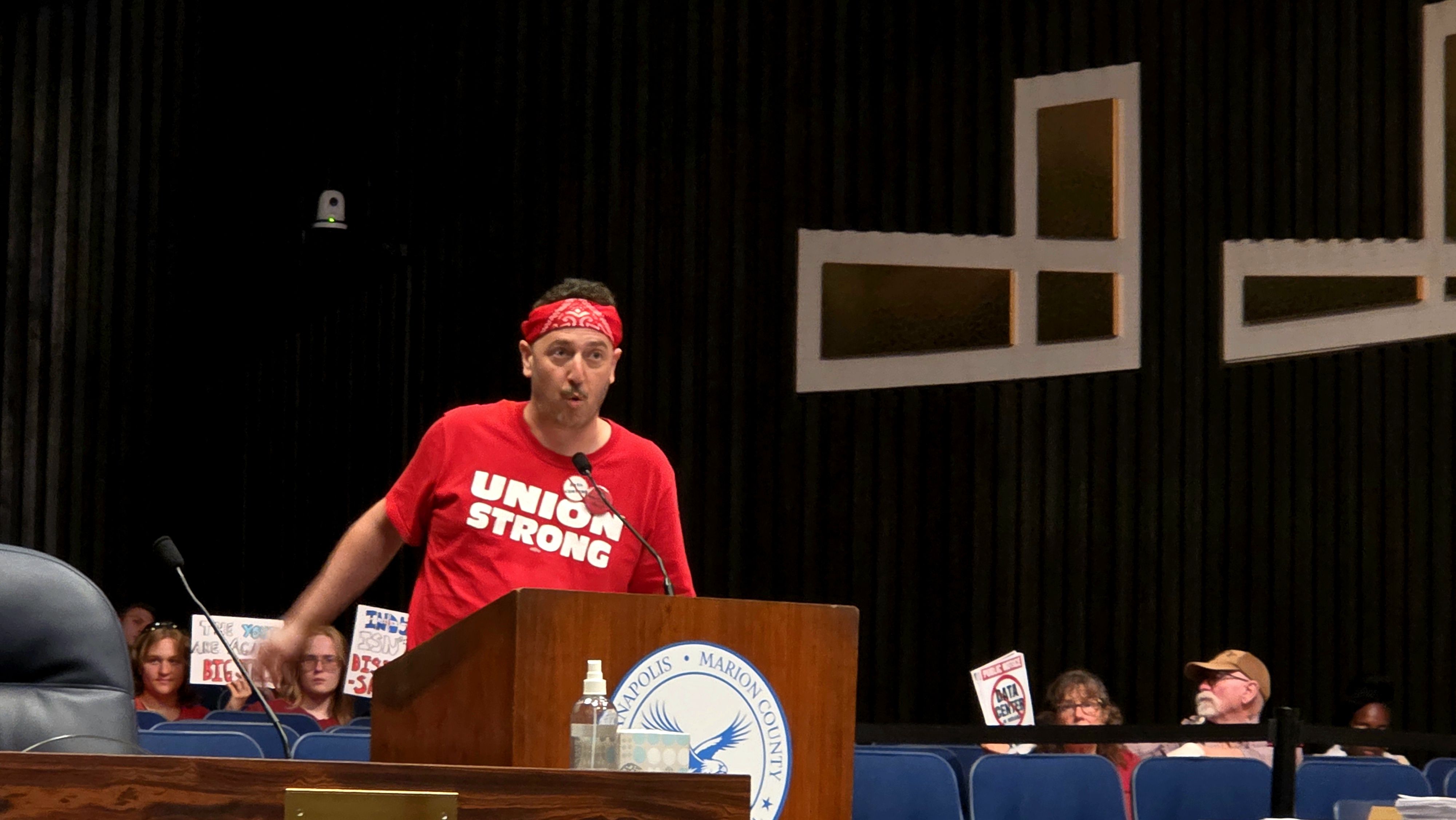 Man in a red bandana and red shirt reading "UNION STRONG" speaks at a podium on a stage with a dark striped wall; audience sits behind, and large white geometric signage decorates the backdrop.
