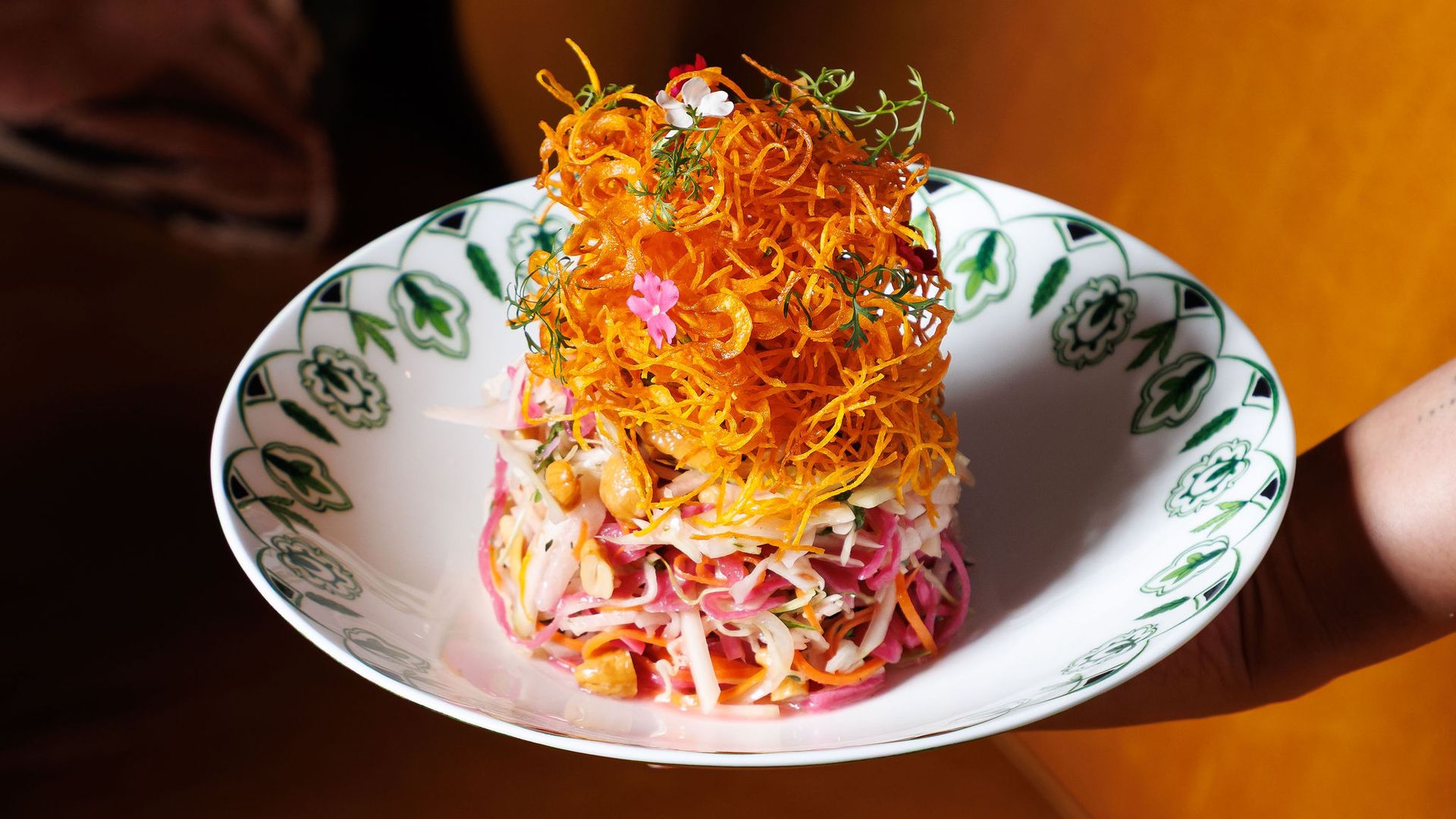 Tall nest of crispy orange fried noodles sits atop a carrot and beet slaw, garnished with herbs and tiny flowers, on a white plate with green floral edge, held by a hand.