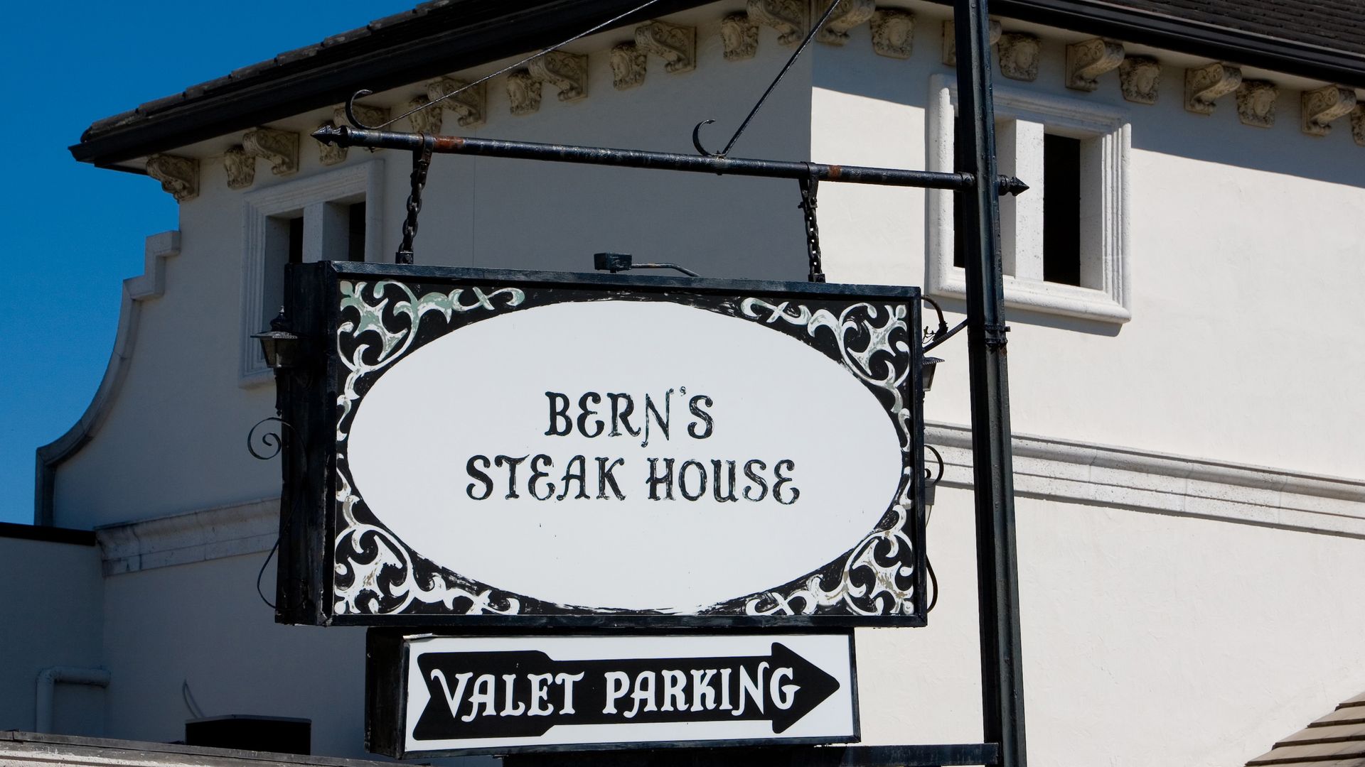 Black and white decorative sign for Bern's Steak House with a valet parking arrow, mounted on a black pole with a white building and clear blue sky in the background.