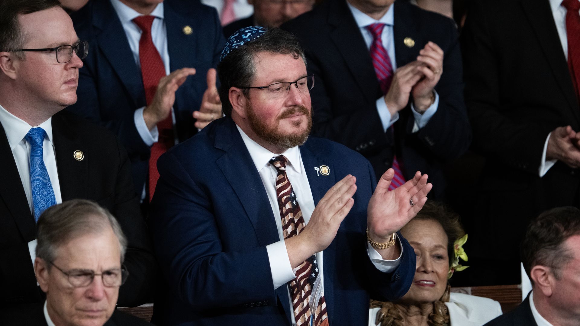 Group of people in formal business attire applauding; center features a bearded man with glasses in a blue suit, patterned tie, and blue kippah, surrounded by others.