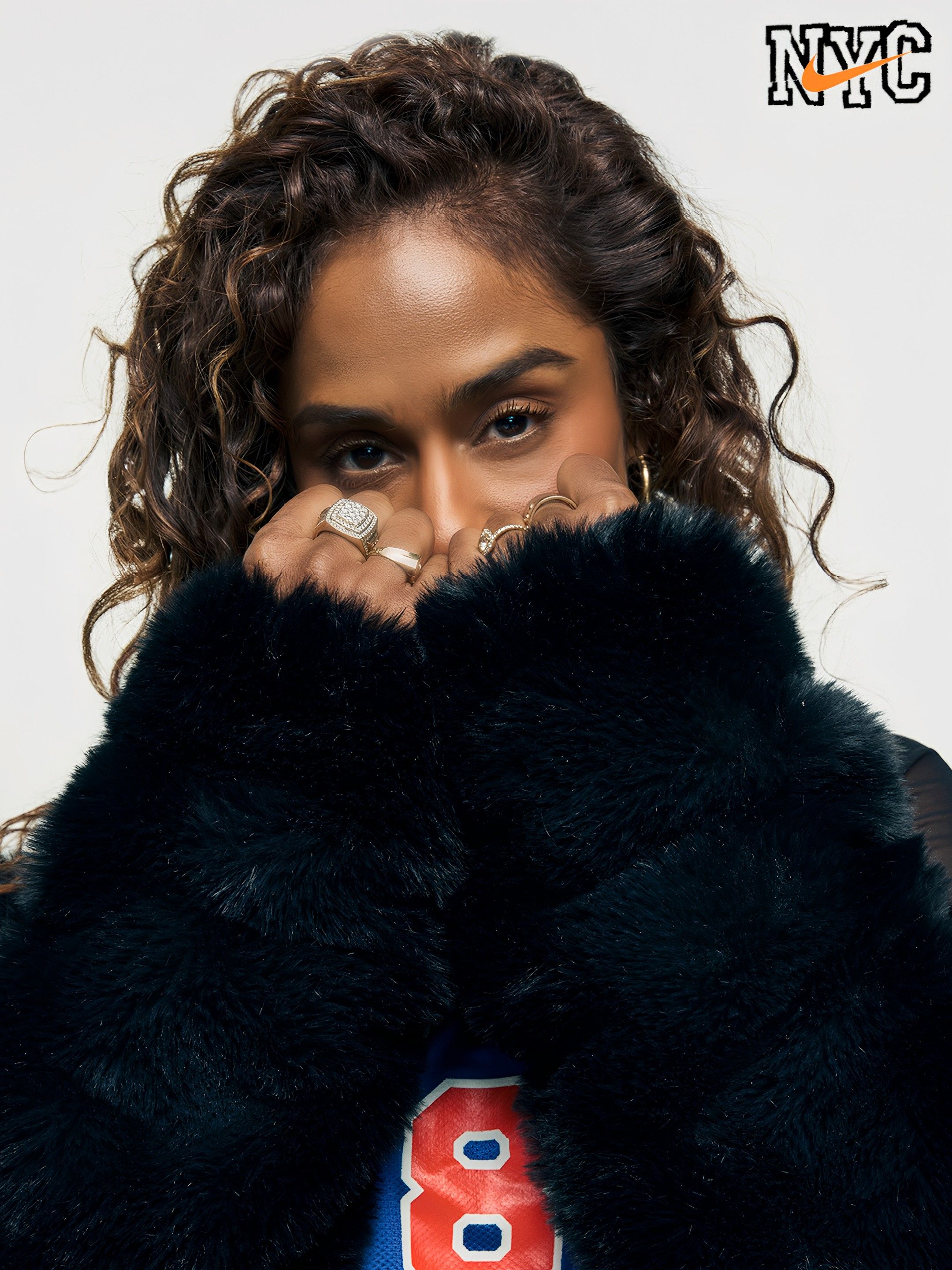 Close-up of a person with curly dark hair, partially hiding behind a black fur coat and wearing multiple rings, eyes visible. NYC logo with an orange Nike swoosh in the top right.