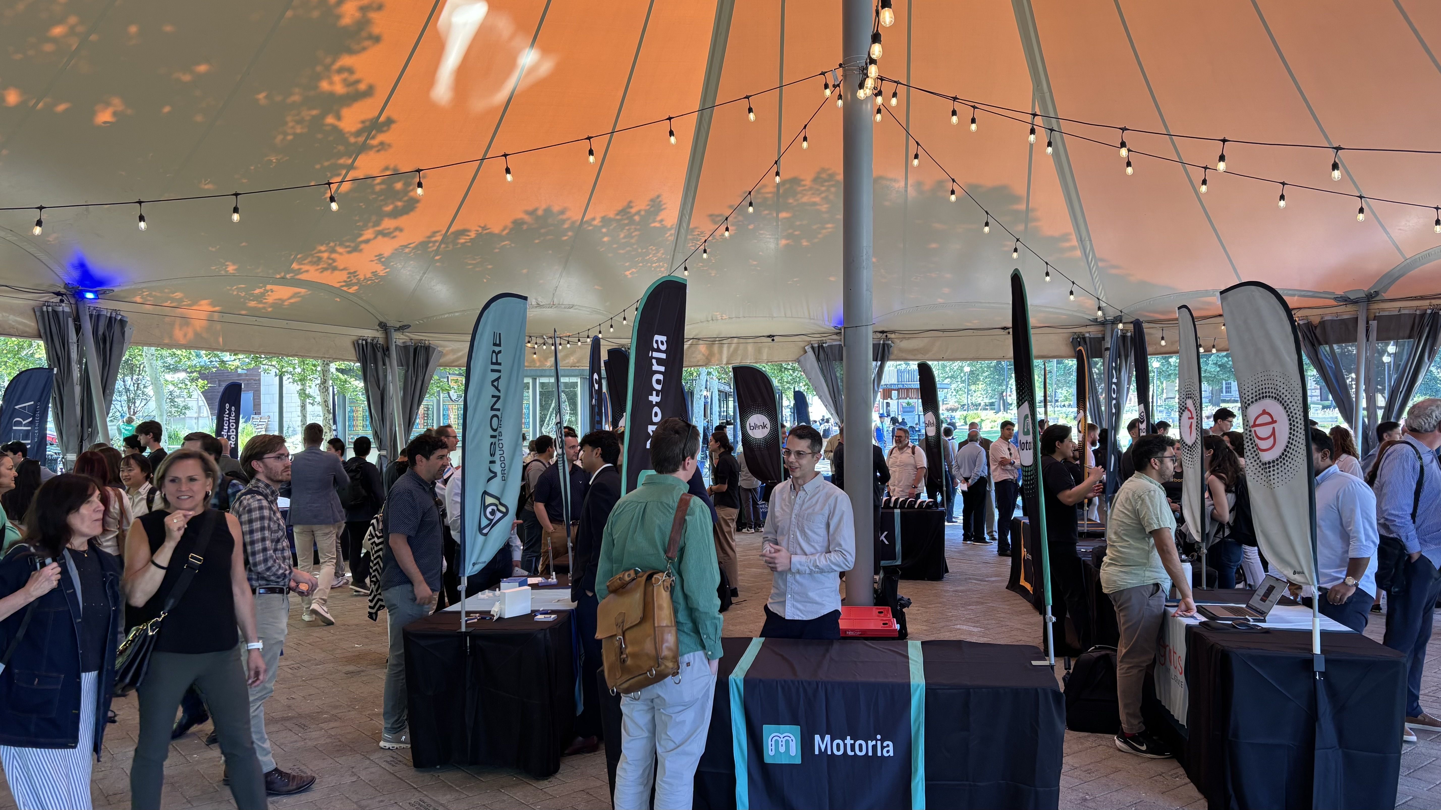 Busy expo under a large orange tent with string lights. Crowds gather around booths with blue and black banners, tables and laptops, creating a lively conference-like atmosphere.