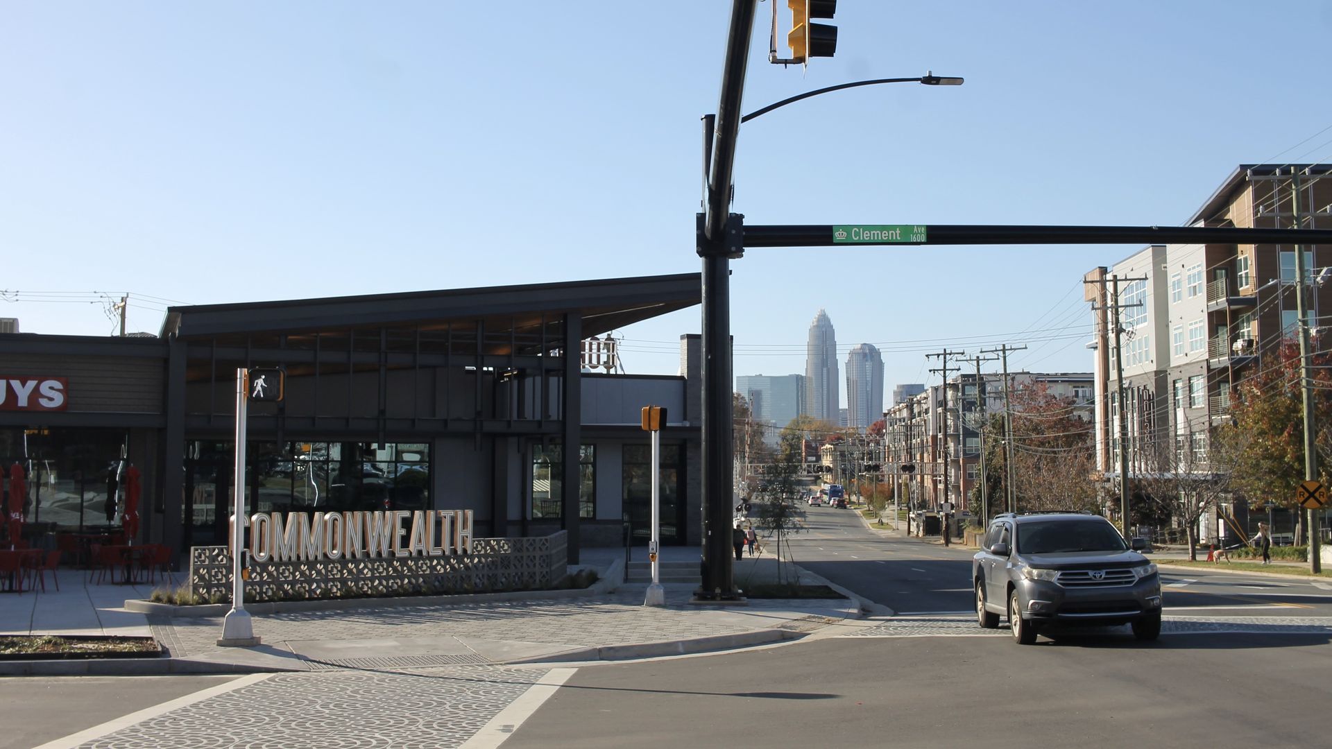 Urban street intersection with traffic lights, a car, and a building with a sign reading "COMMONWEALTH." Clement Ave street sign is visible with a clear blue sky and city skyline in the background.