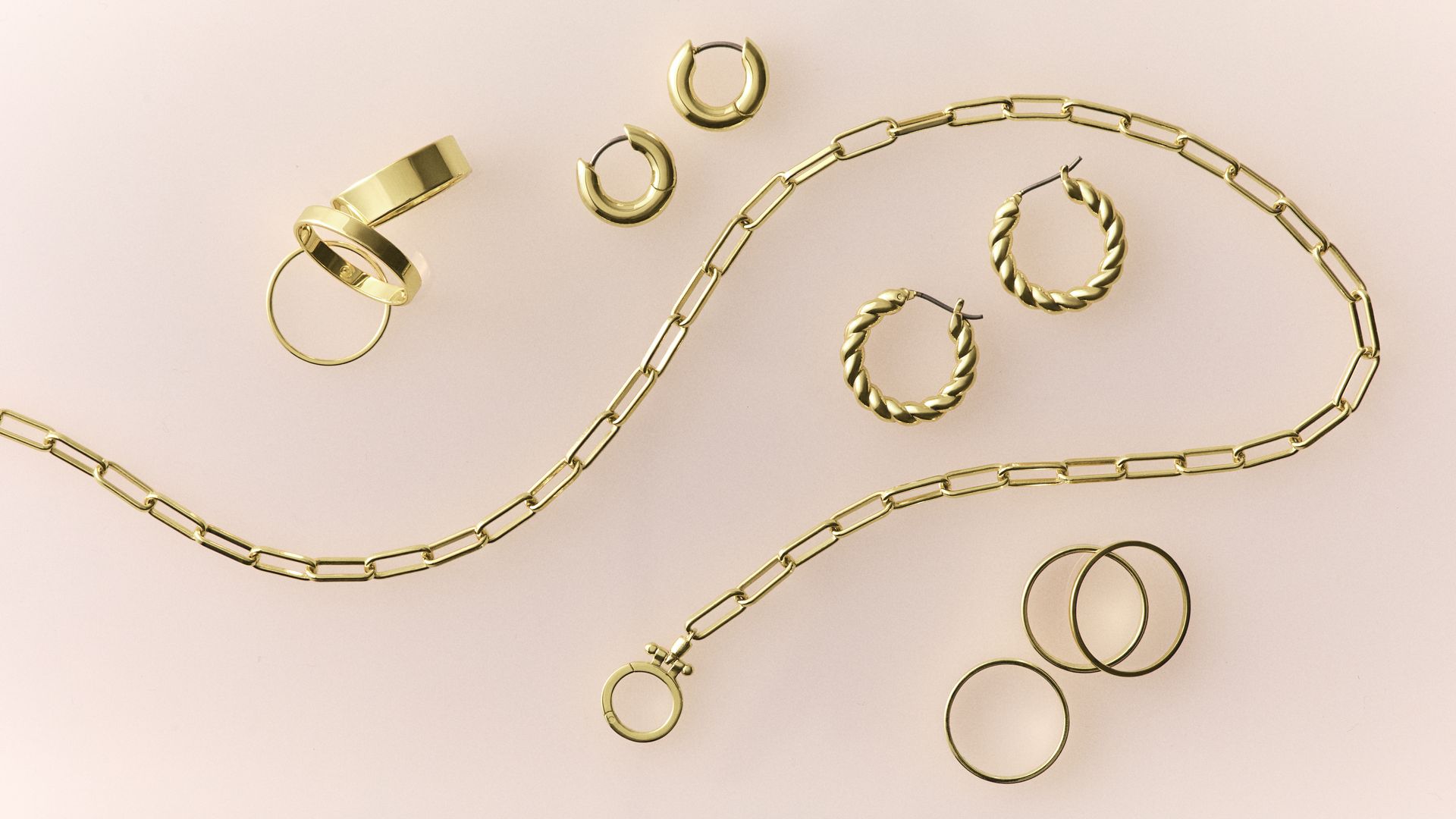 Various gold jewelry pieces on pale pink background, including a chain necklace, twisted hoop earrings, smooth hoop earrings, and multiple simple gold rings.