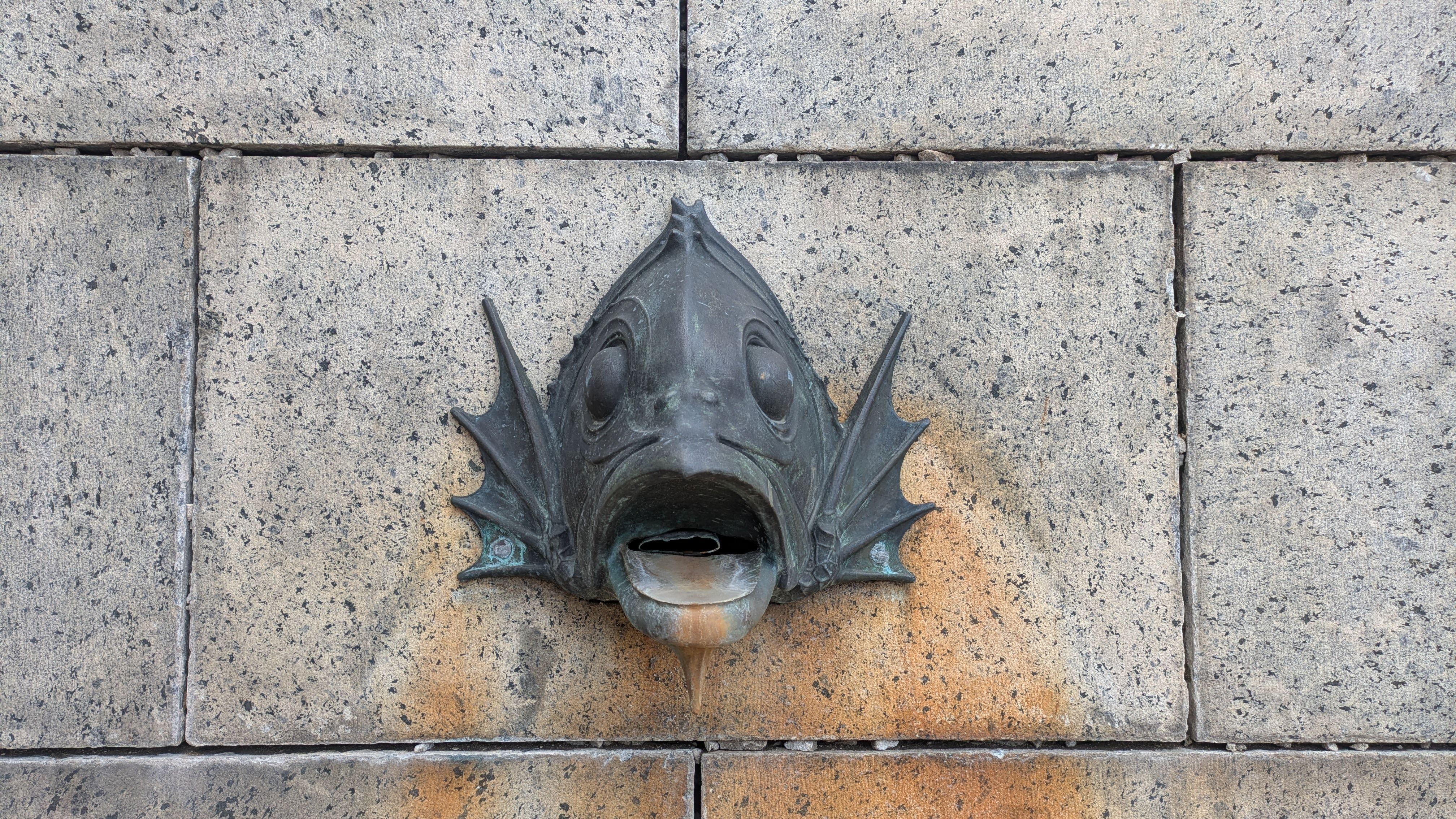 Bronze fish-head water spout mounted on a gray speckled brick wall; its open mouth acts as the spout, with large eyes and decorative fins on either side, and orange rust staining beneath.