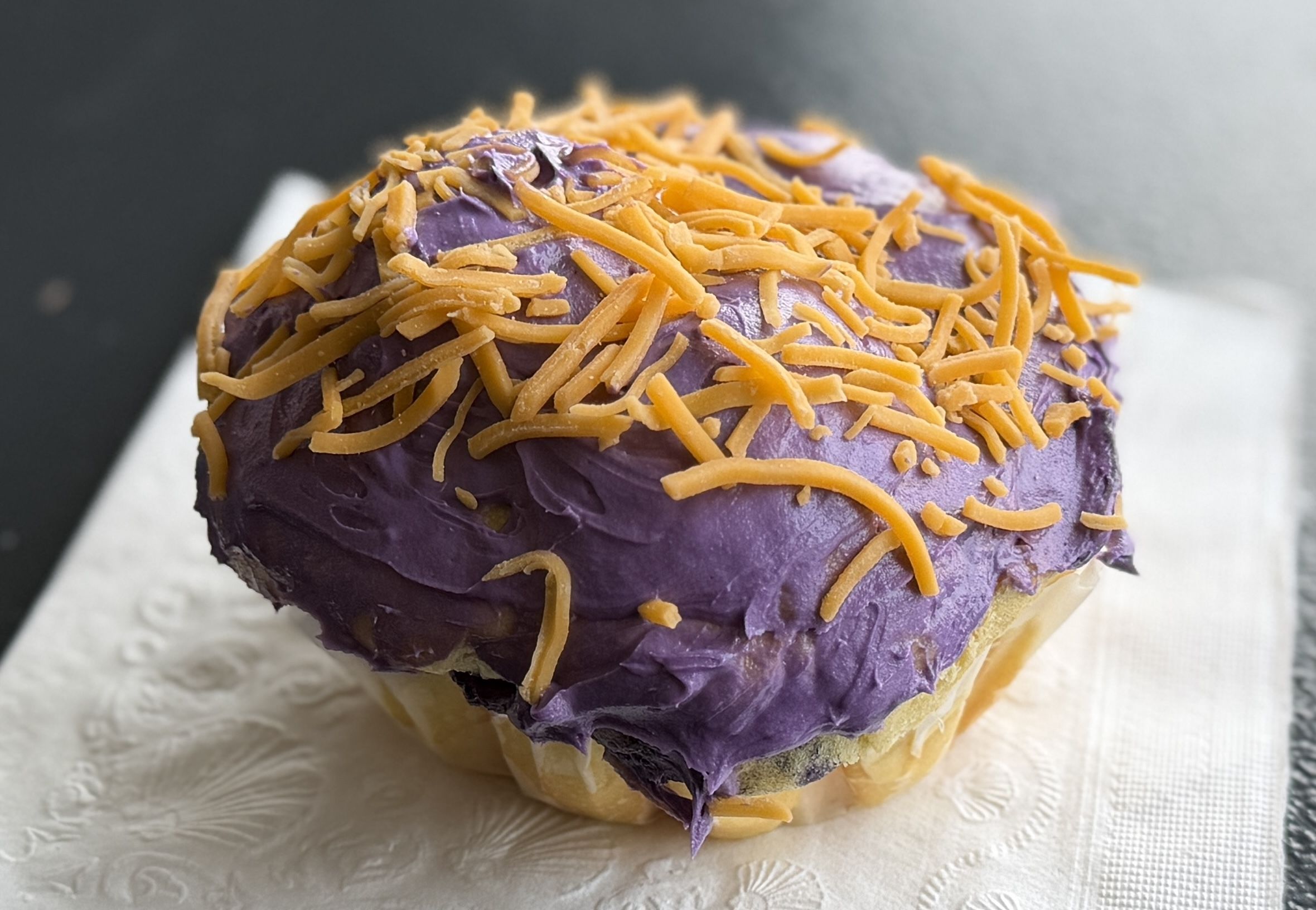 Close-up of a doughnut with purple frosting and orange shredded topping, resting on a white patterned napkin against a dark, blurred background.