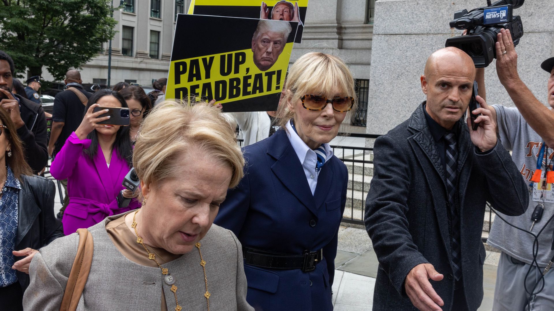 Writer E. Jean Carroll, wearing bleached blonde hair tied back, tortoiseshell tinted brown glasses, a navy coat and blue blouse,  leaves the courthouse in September 2024 in New York City as a protesters behind her holds a placard of Trump saying "Pay up, Deadbeat."