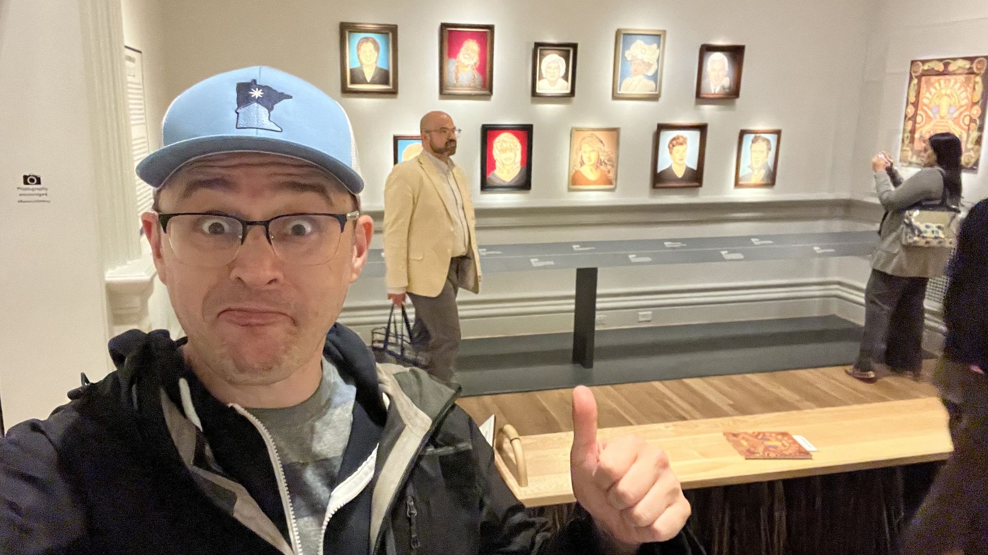Man in a blue cap and glasses giving a thumbs-up inside an art gallery with portraits on the wall behind him, a man in a beige jacket walking, and a woman taking a photo.
