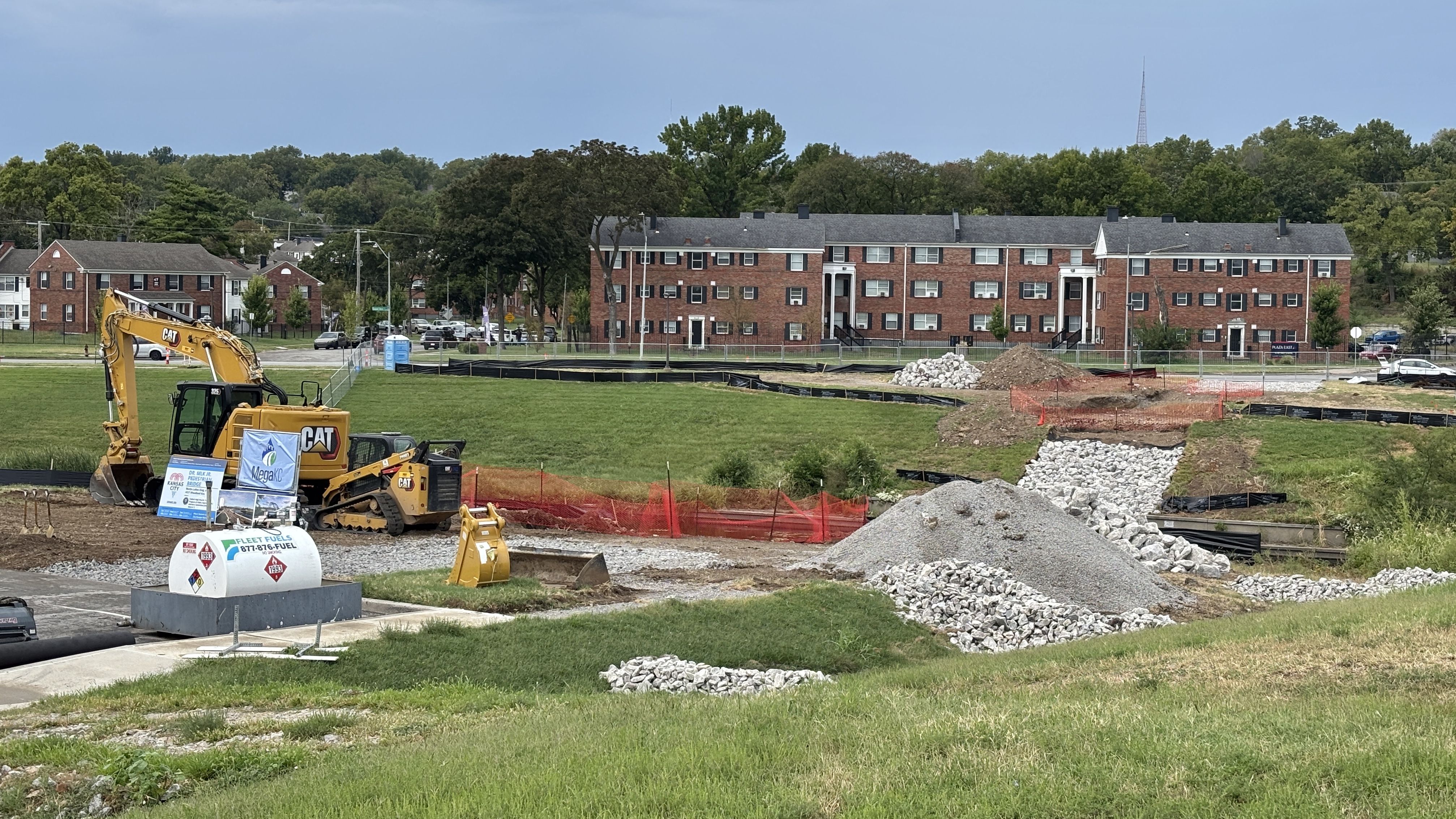 Construction site in the foreground with a yellow CAT excavator, dirt piles, and orange safety fencing; a line of red brick apartment buildings and trees sit in the background under a blue sky.