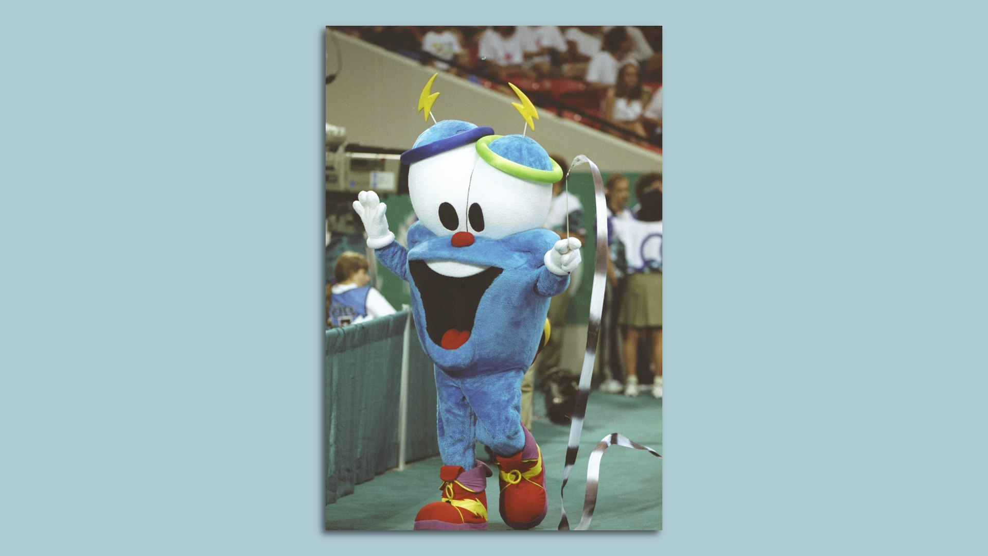 A photo of a blue mascot with a big smile and eyes and lightning bolts for eyebrows waves a streamer