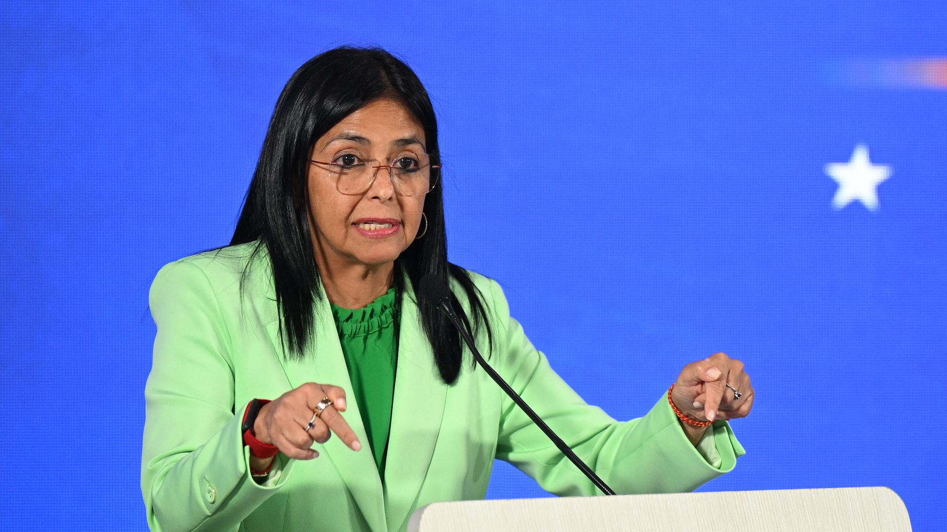 Delcy Rodriguez, wearing a green suit jacket and shirt, speaks into a microphone.