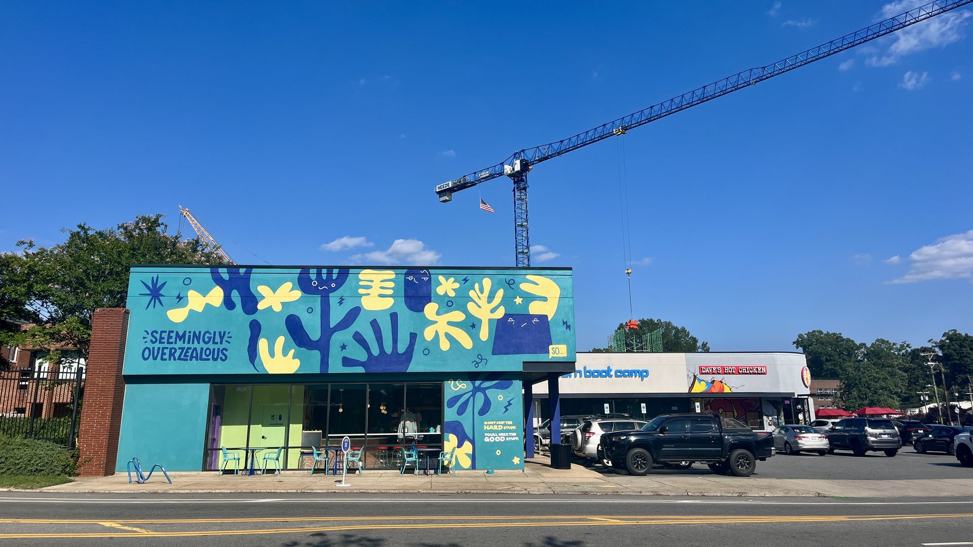 Clear blue sky with a long construction crane diagonally across; below is a turquoise storefront mural with yellow and blue abstract shapes, outdoor chairs, and a row of parked cars.
