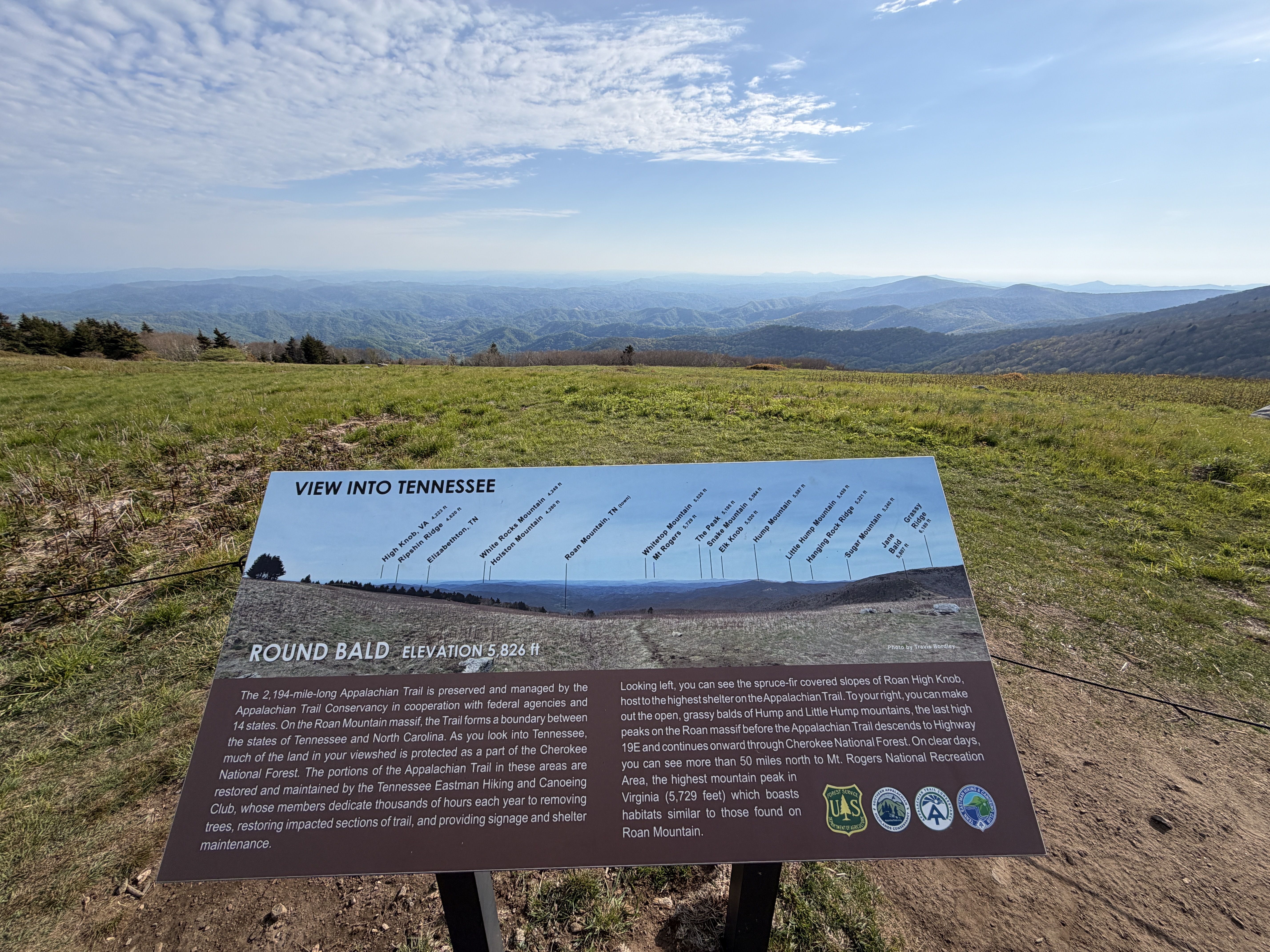 Panoramic mountain vista from a grassy overlook. A large sign in the foreground reads "VIEW INTO TENNESSEE" and "ROUND BALD ELEVATION 5,826 ft." Distant blue ridges roll under a clear, partly cloudy sky.
