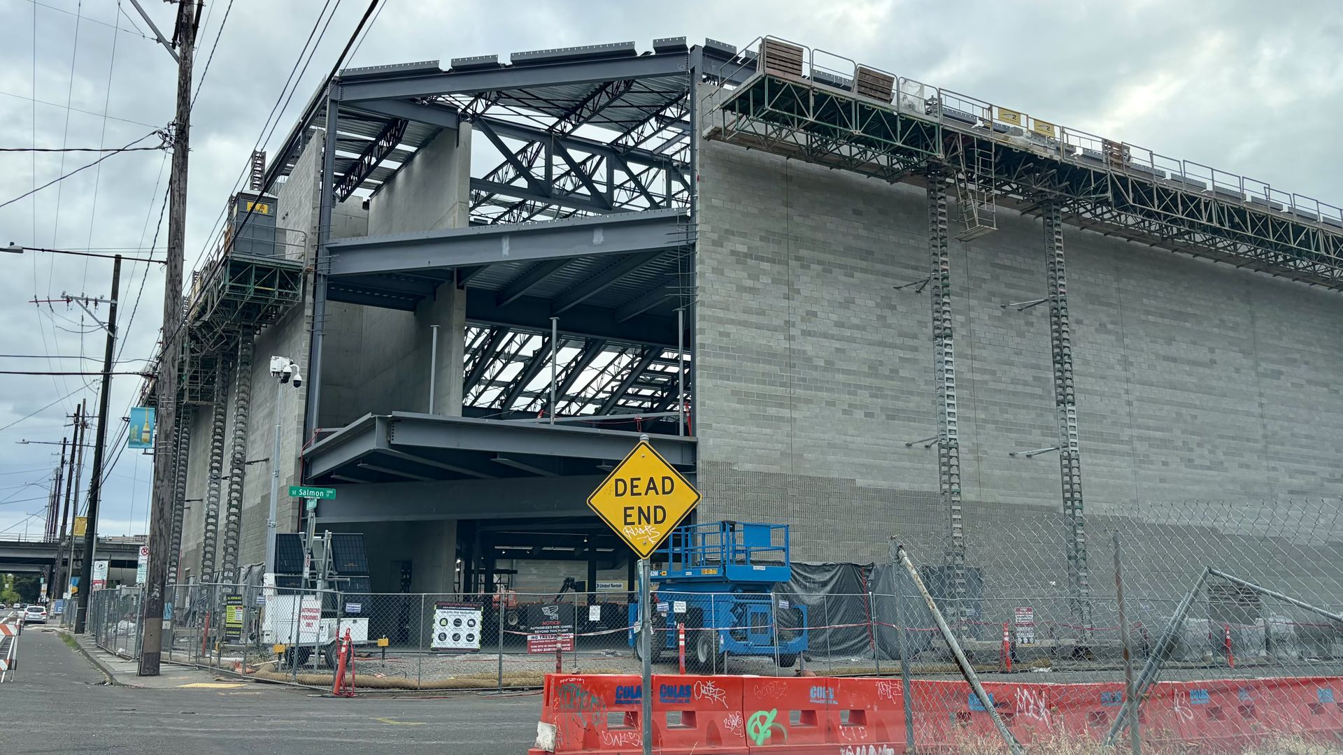 Construction site beneath a steel-framed overpass with gray concrete blocks. Scaffolding and a blue lift by a chain-link fence; a yellow "DEAD END" sign faces the street; orange barriers with graffiti; cloudy sky.