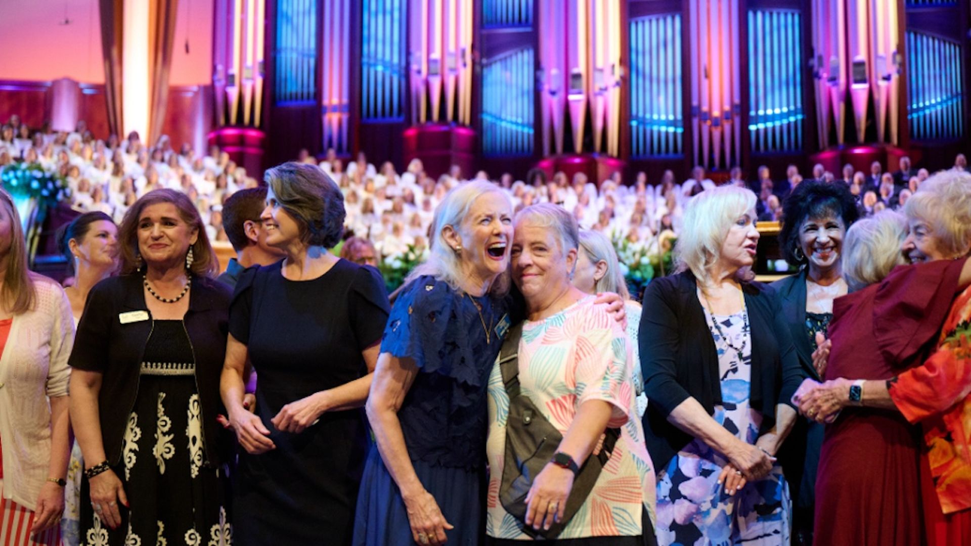 A group of women, two of whom are hugging, stand in front of a large choir.