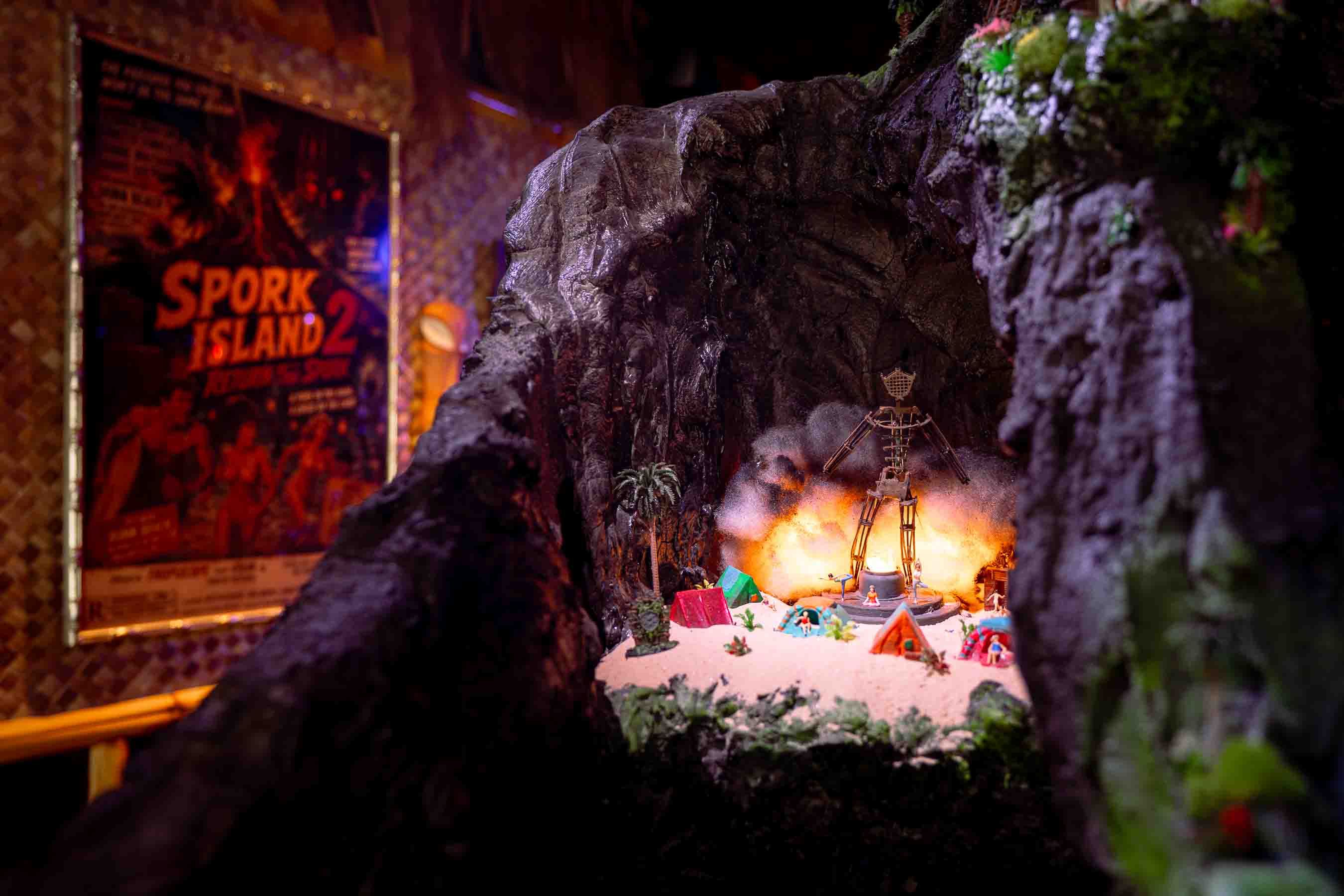 Nighttime diorama in a rocky alcove: a sandy crater with tiny tents and palm trees around a campfire, a skeletal tower nearby, and a left-wall poster reading "SPORK ISLAND 2".