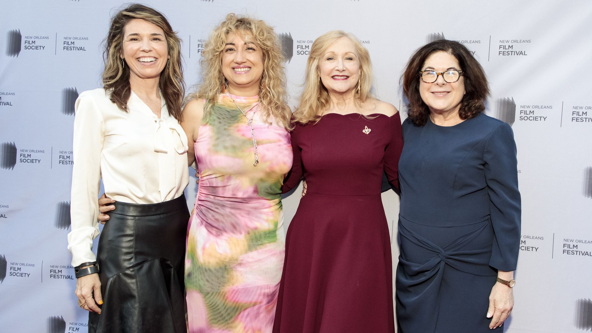 Four women in cocktail attire hold their arms around each other and smile on a red carpet.