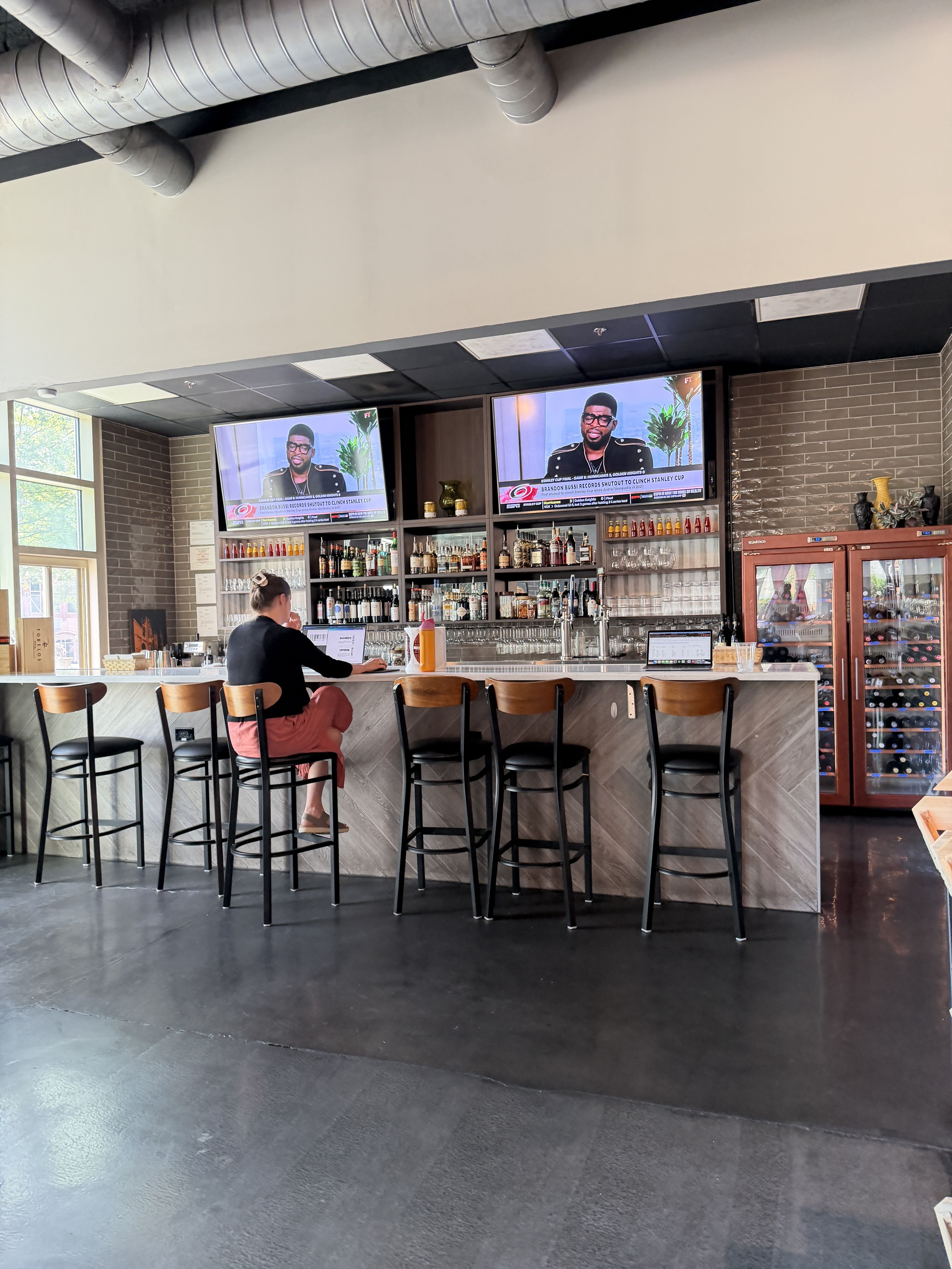 Bar interior with a long counter and black stools; liquor shelves behind; two TVs show a man; a person sits at the counter using a laptop; brick walls and exposed silver ducts above.