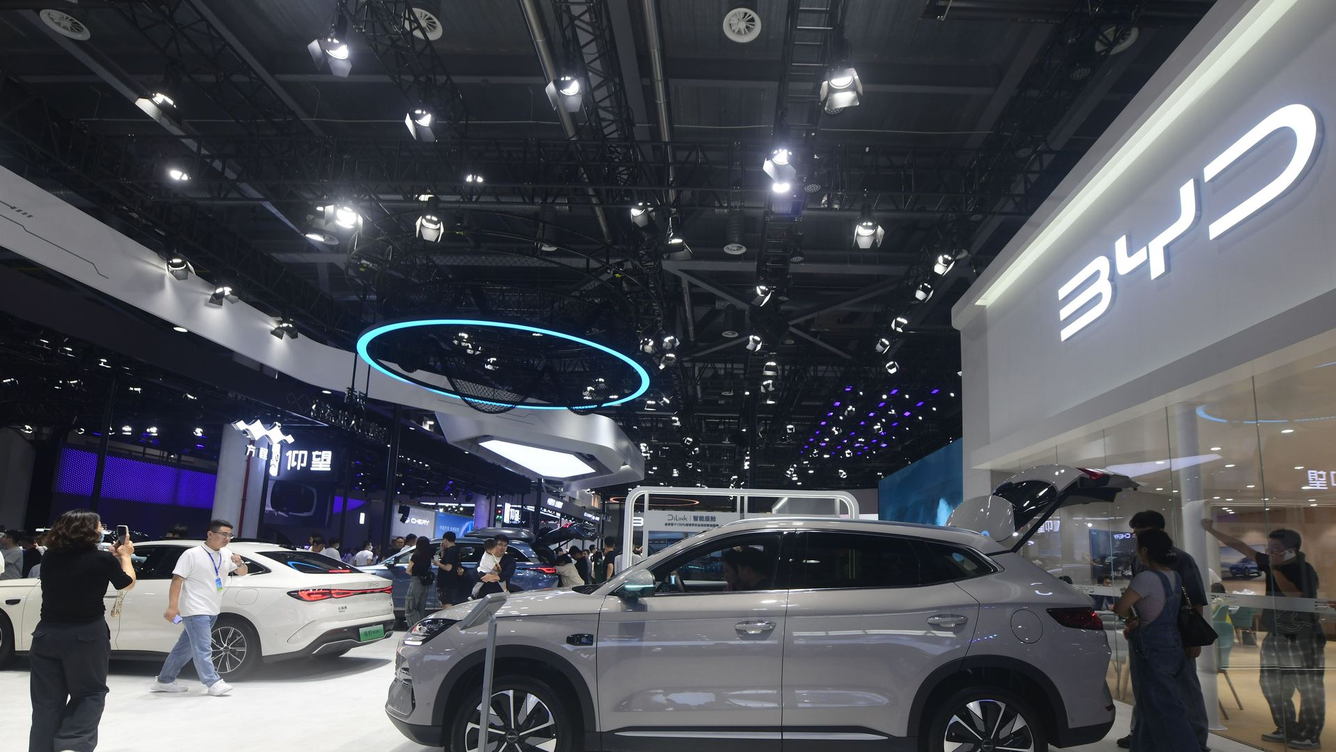 Gray BYD electric SUV displayed in a bright showroom with people around, a white sedan nearby, under ceiling lights and a blue circular light fixture.