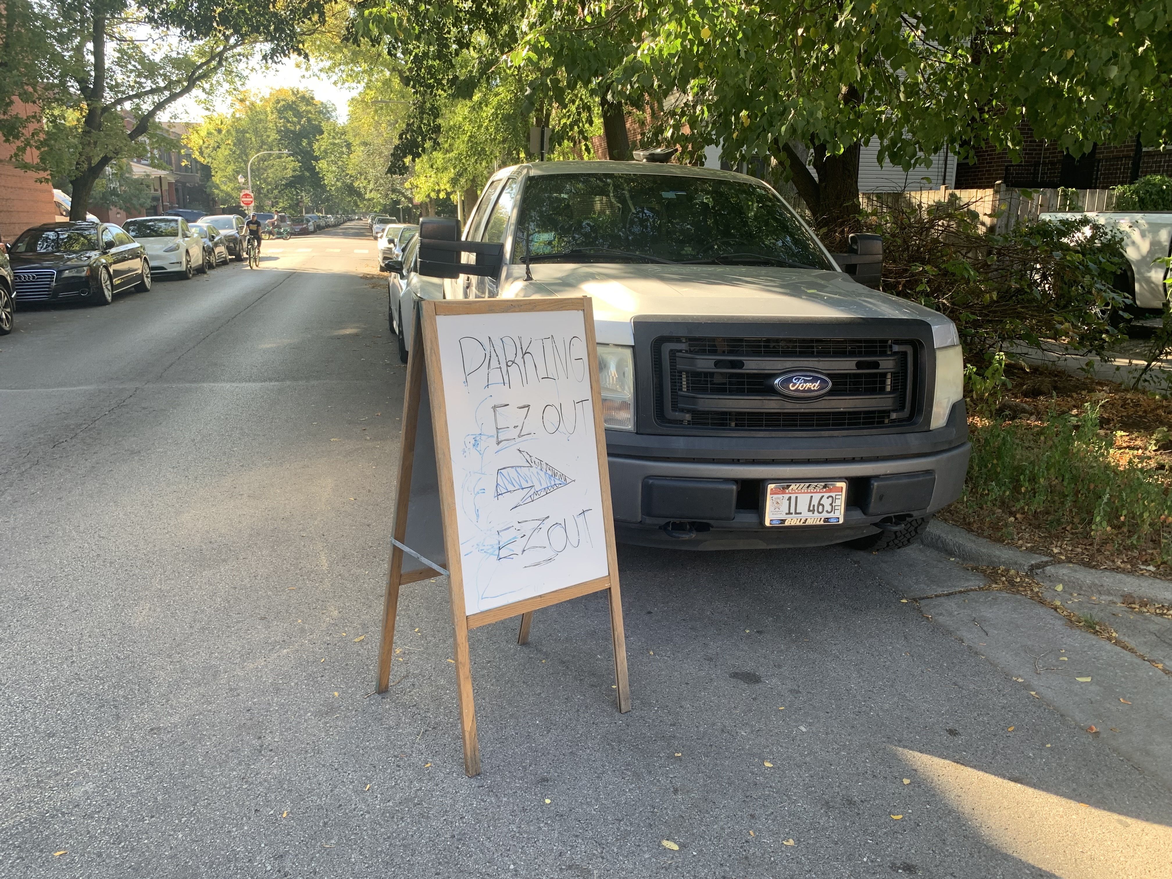 A silver Ford truck parked on a tree-lined street beside a wooden sandwich board with handwritten text "PARKING E.Z. OUT" and an arrow pointing right, directing parking exit.