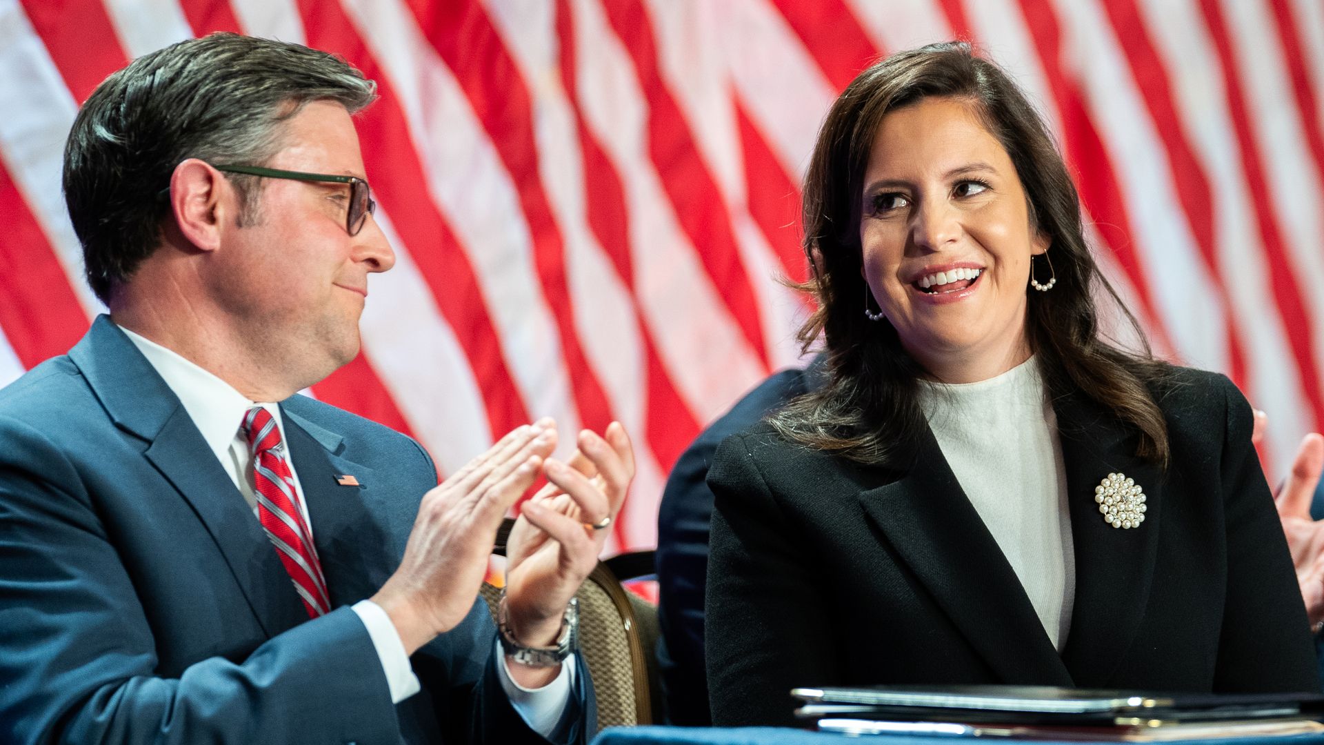 Speaker Mike Johnson, wearing a blue suit, and Rep. Elise Stefanik, wearing a black suit, sitting in front of a row of American flags.