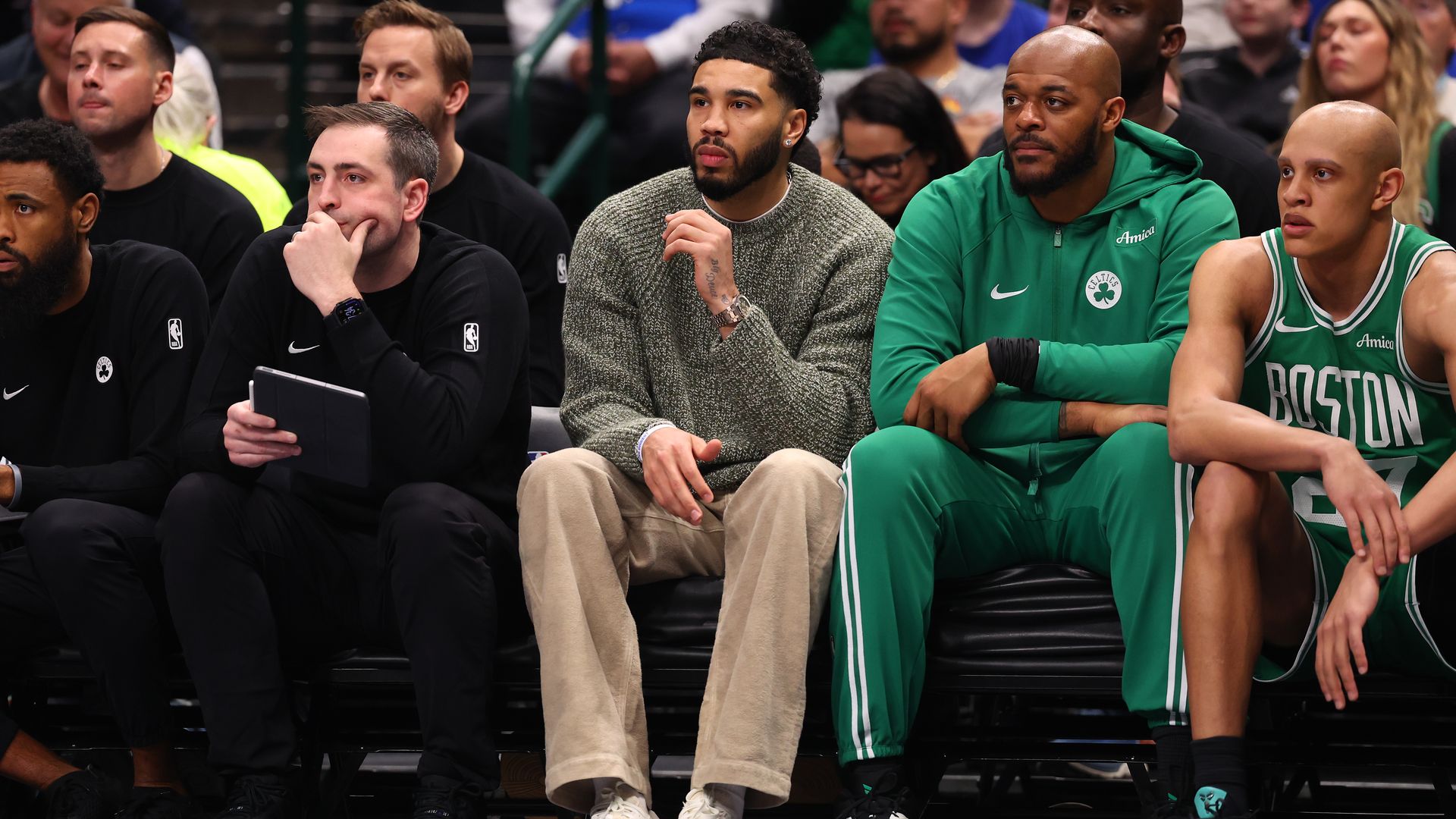Jayson Tatum of the Boston Celtics sits on the team bench during the first quarter against the Dallas Mavericks. Tatum is in street clothes: brown khaki pants and a sweater.