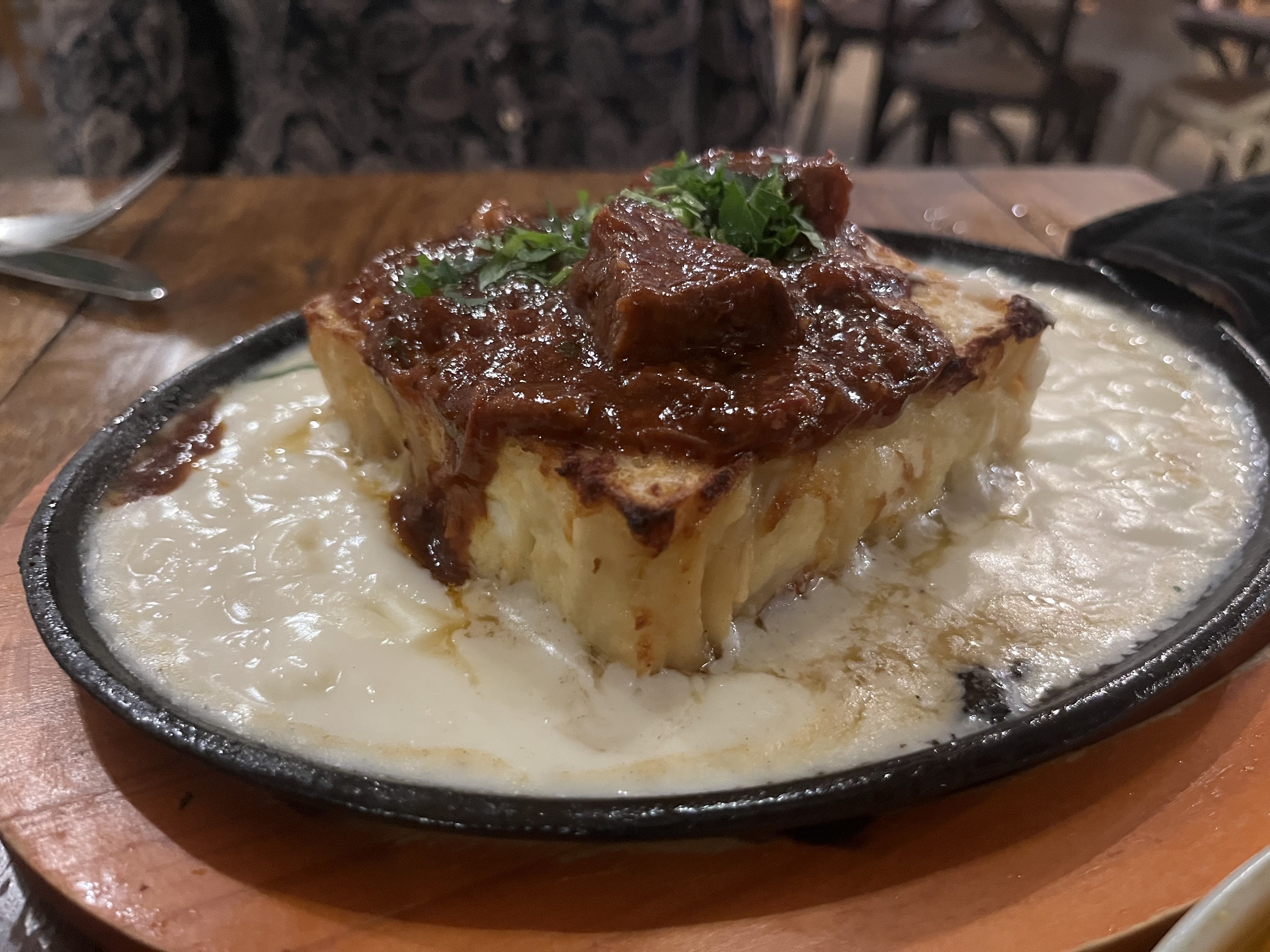 Close-up of a thick slice of toasted bread topped with glossy bone marrow and chopped parsley, served in a small black skillet on a wooden table.