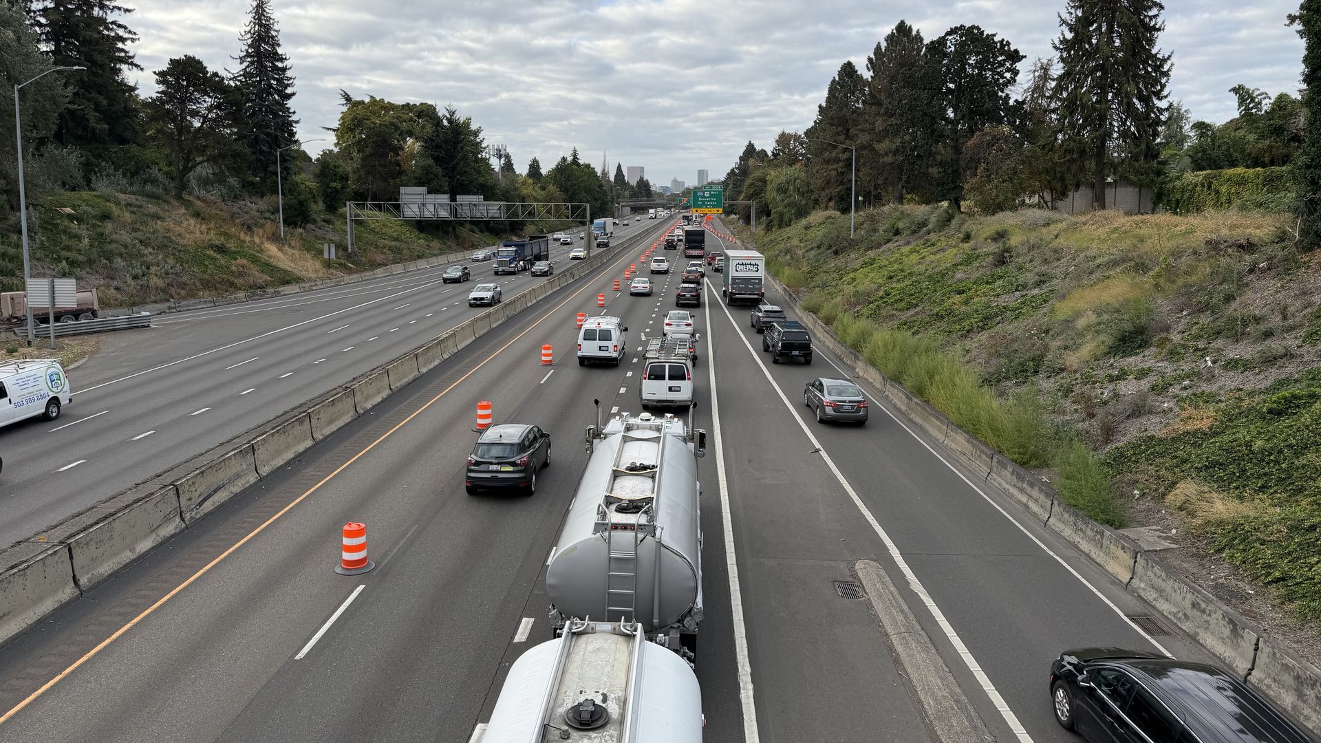Multi-lane highway under a cloudy sky with orange construction cones on the right. A large white tanker truck leads the foreground as cars travel in both directions, bordered by trees and greenery.