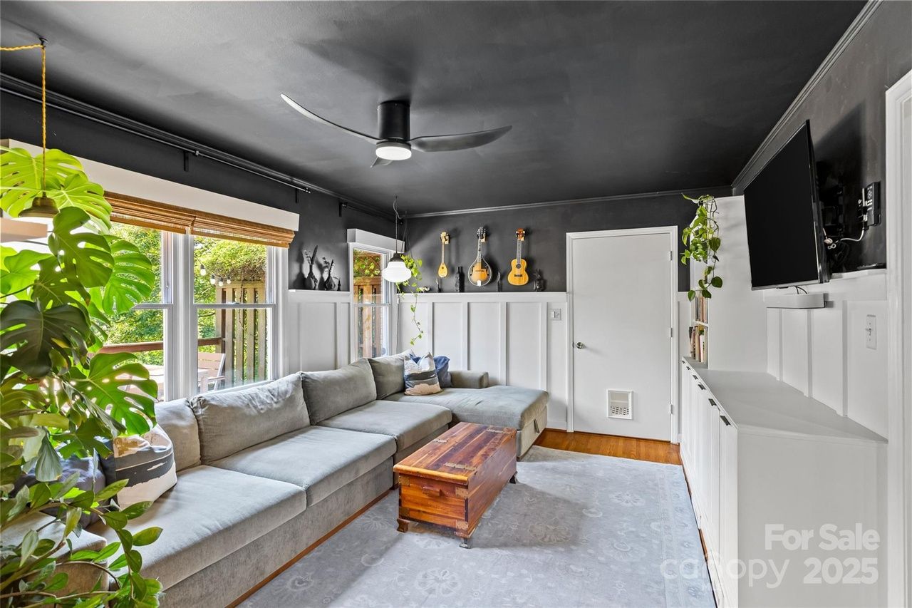 Modern living room with dark ceiling, light gray sectional sofa, wooden coffee table, potted plants, guitar wall mounts, white wainscoting, flat-screen TV, and large windows with views of greenery.