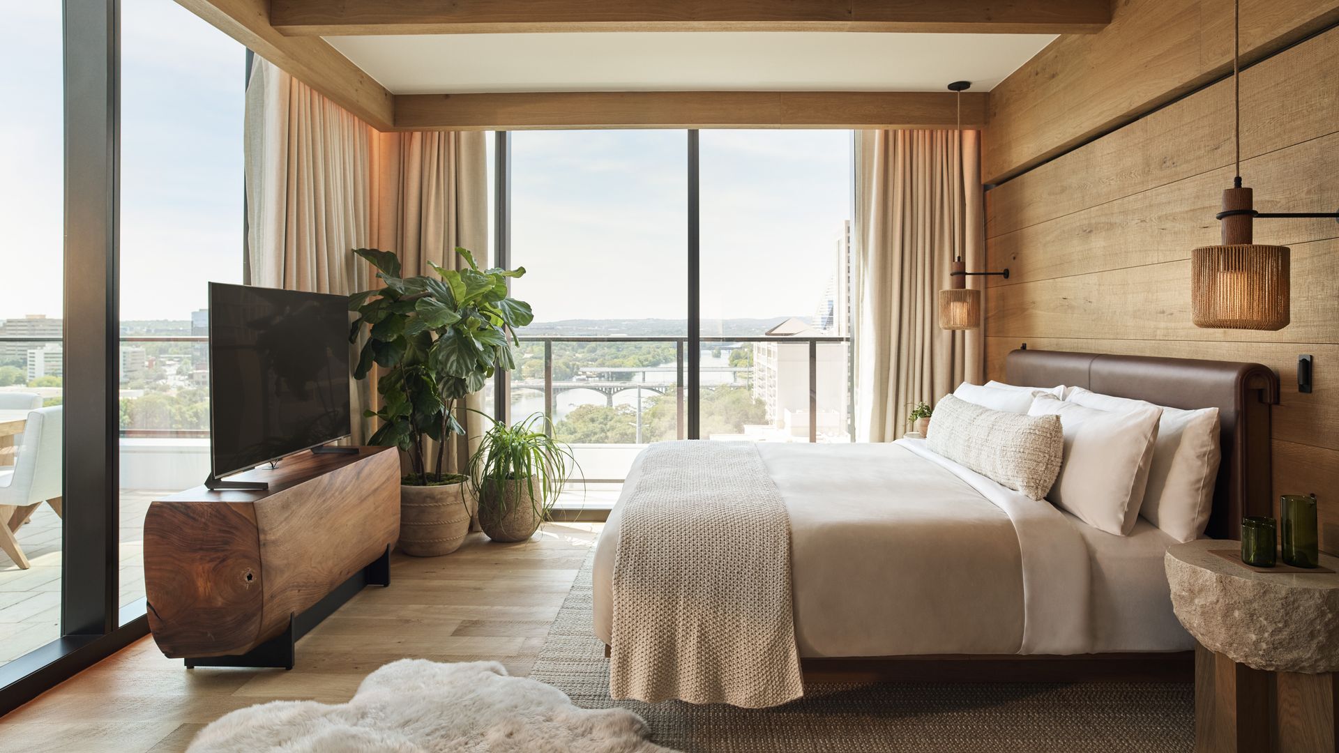 Bright bedroom with warm wood beams, floor-to-ceiling windows with beige curtains, a low bed in cream bedding, pendant lights, plants, and a TV on a wooden stand; city skyline outside.