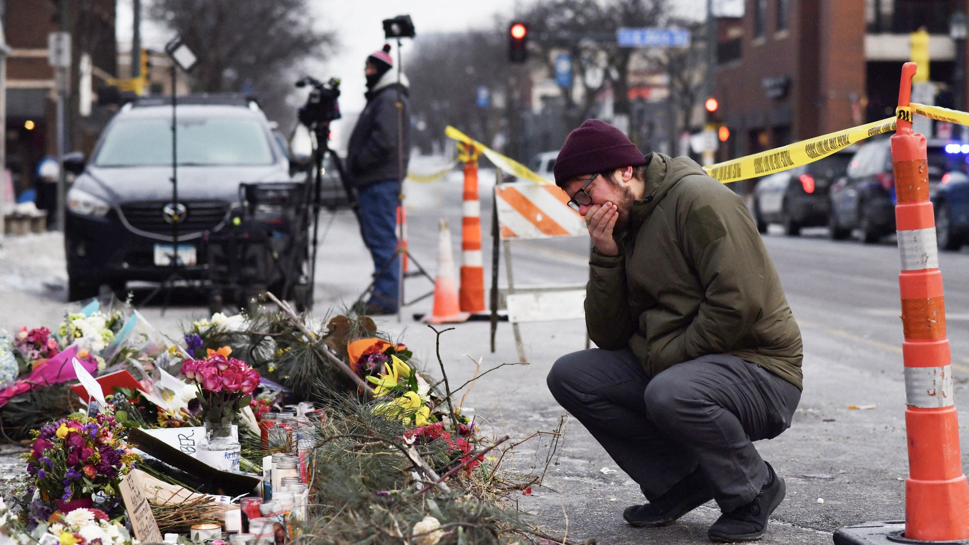 A person in a red beanie, glasses, gray pants and an army green jacket crouches down with his hand over his mouth by a vigil in the street.