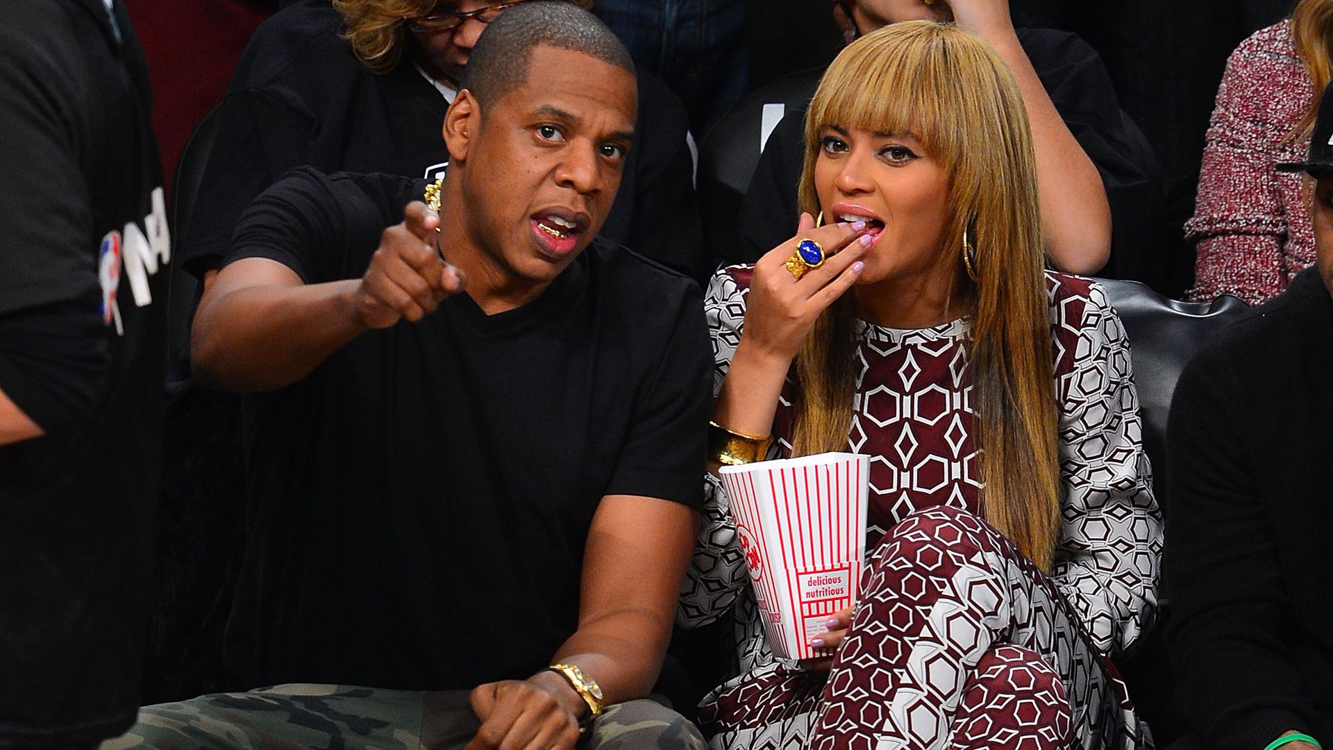 Beyonce speaks with Jay-Z, courtside as a basketball game. Beyonce is eating popcorn, and Jay-Z points toward the game.