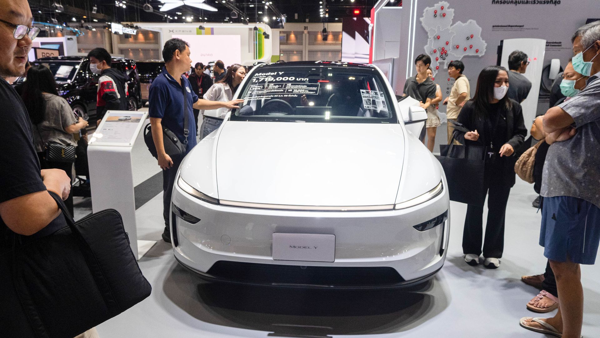 White Model Y electric car on display at a Tesla booth with people around, showing price and features on windshield, inside a brightly lit exhibition hall.