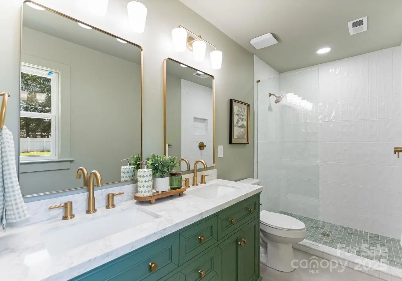 Modern bathroom with green vanity, white marble countertop, gold fixtures, two mirrors, potted plants, large walk-in shower with white tiles, and a toilet.