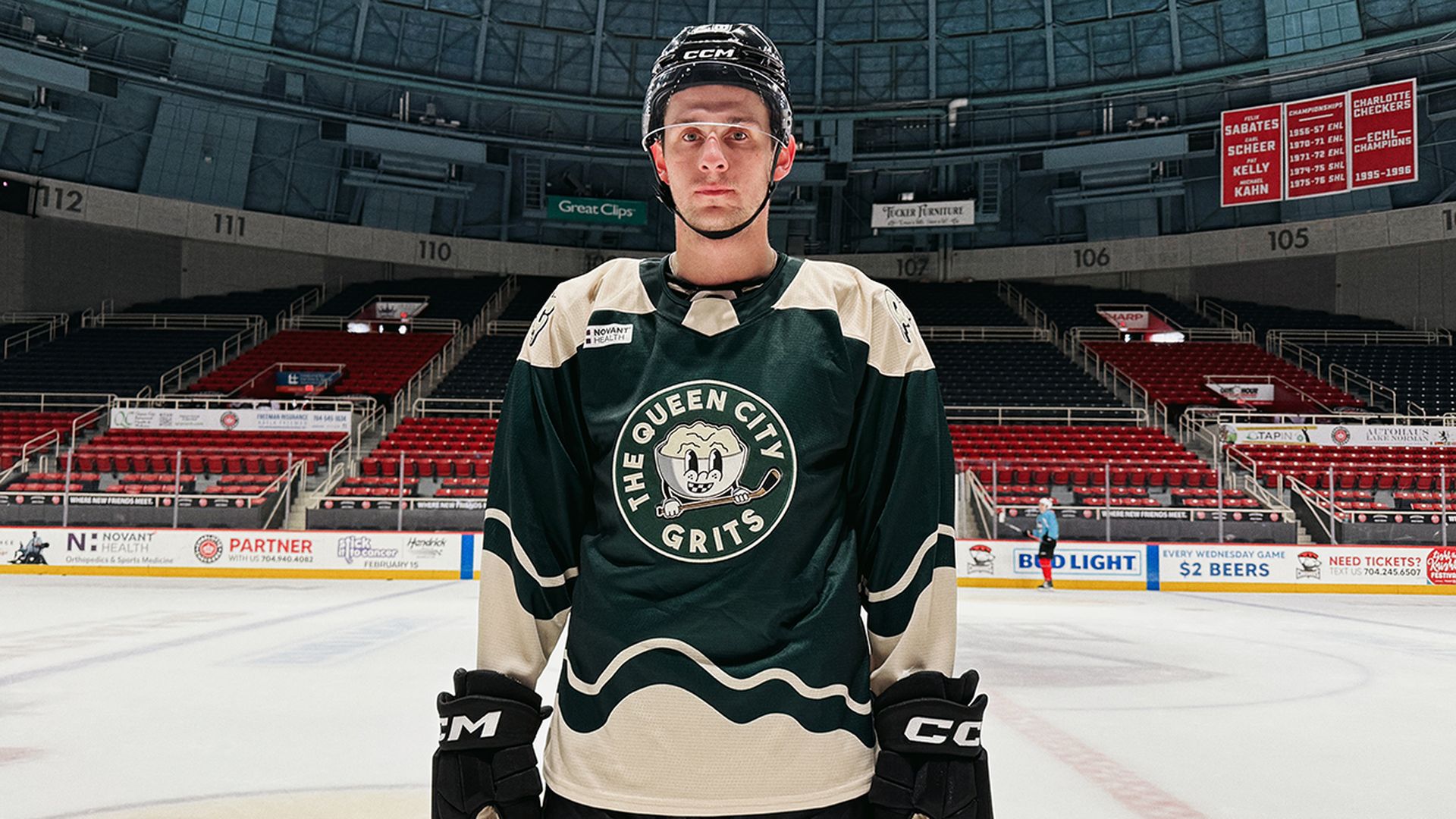 Hockey player in black and beige The Queen City Grits jersey and helmet stands on ice rink with empty red and black seats in background.