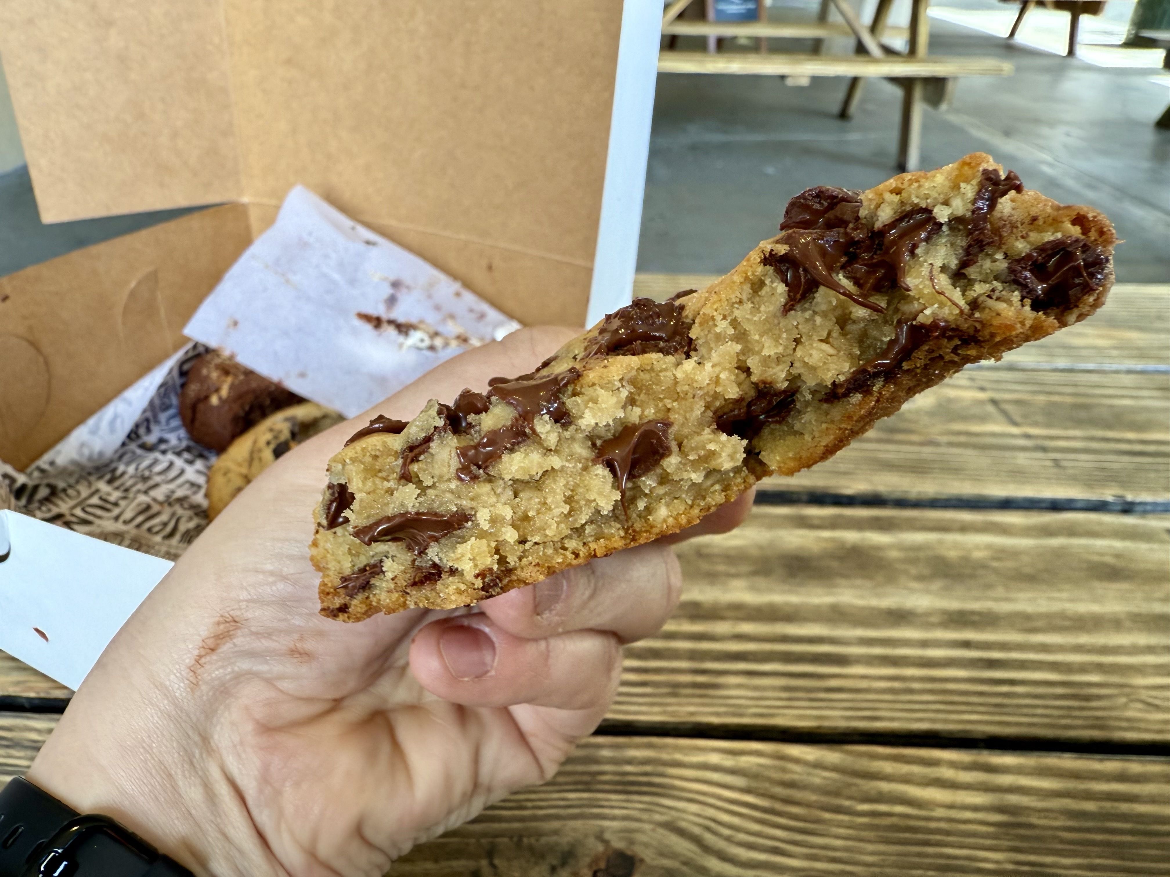 Hand holding a thick, golden-brown chocolate chip cookie with melted chocolate chunks over a wooden picnic table, with more cookies in a brown box in the background.