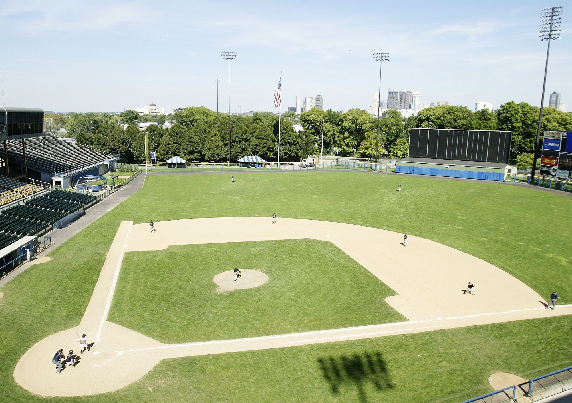 A baseball game in progress at Cooper Stadium with players in black and white uniforms, empty bleachers, tall light poles, American flag, and city skyline with trees in the background under a clear sky.